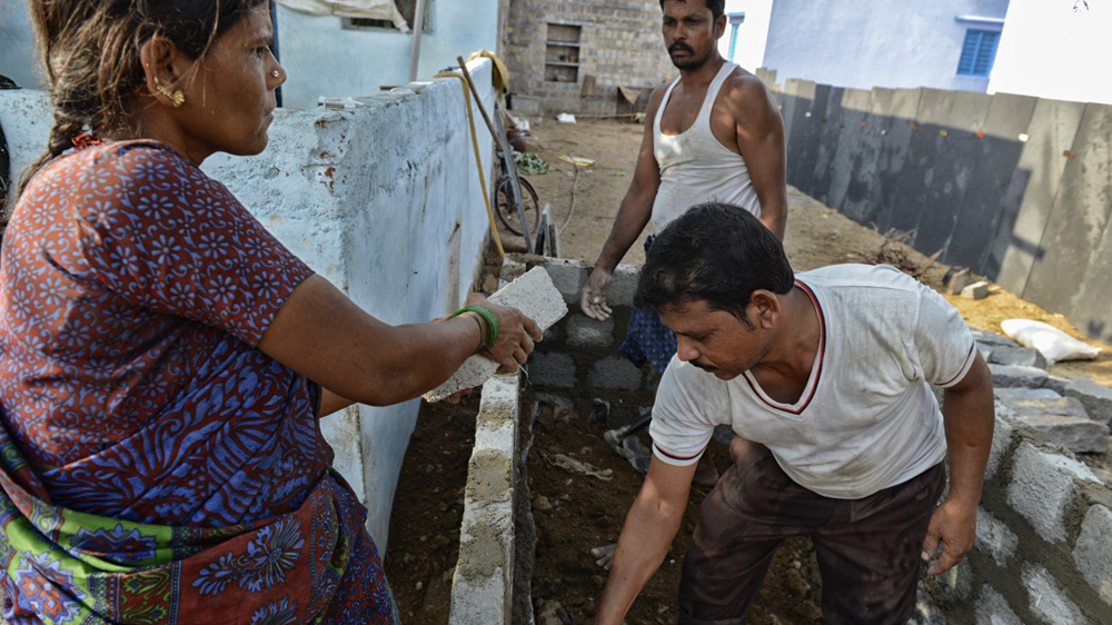 Savithramma Sugali passes a brick to a worker hired to build their toilet as her husband, Rangudunaik, looks on in Pendlimanu village [Zigor Aldama/Al Jazeera] 