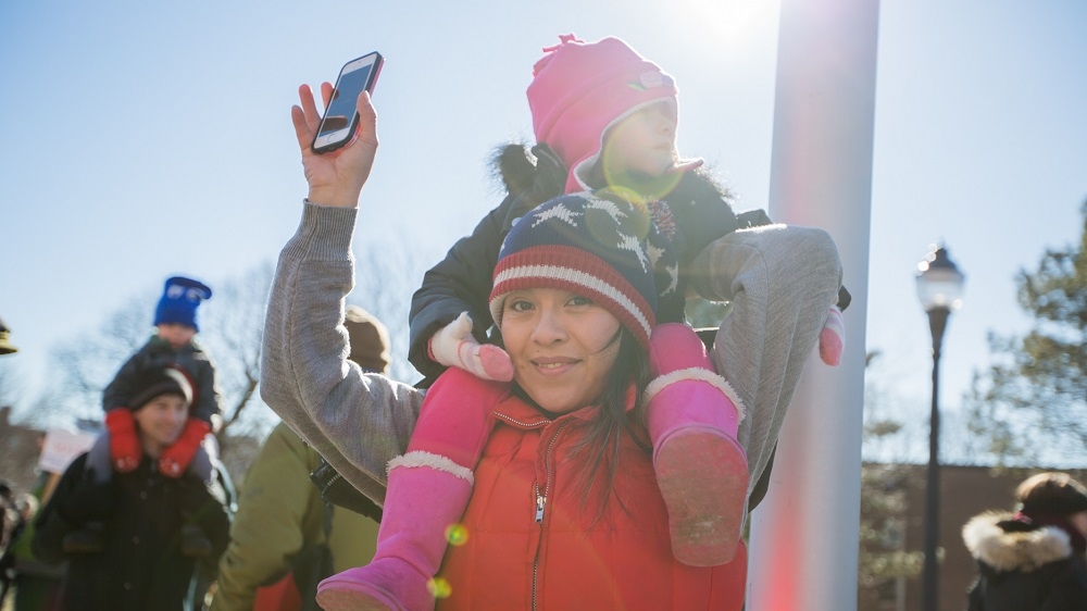 Sandra Suarez holds her daughter Enola on her shoulders, as they listen to music, during the We Are One rally outside Somerville High School [Carolyn Bick/Al Jazeera]