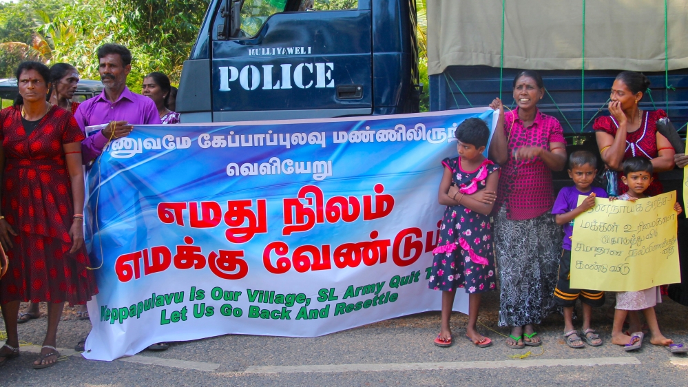 Protesters stand in front of a police truck that has been parked across the road from the gate, with a banner that reads "We want our land back" in Tamil [Lisa Fuller/Al Jazeera]