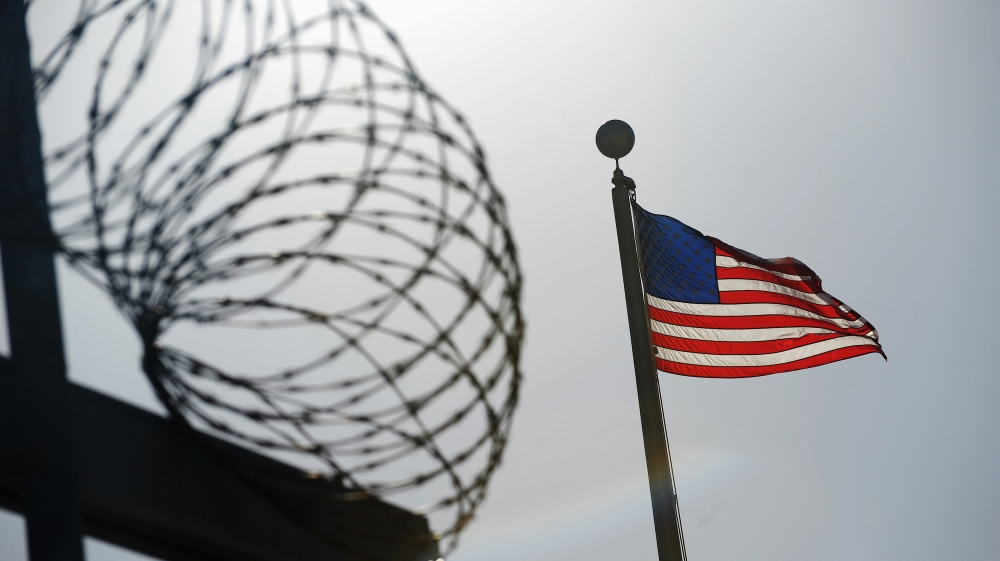 Barbed wire with the US flag to the right of it. The wire and fence is black, like a silhouette against the sky.