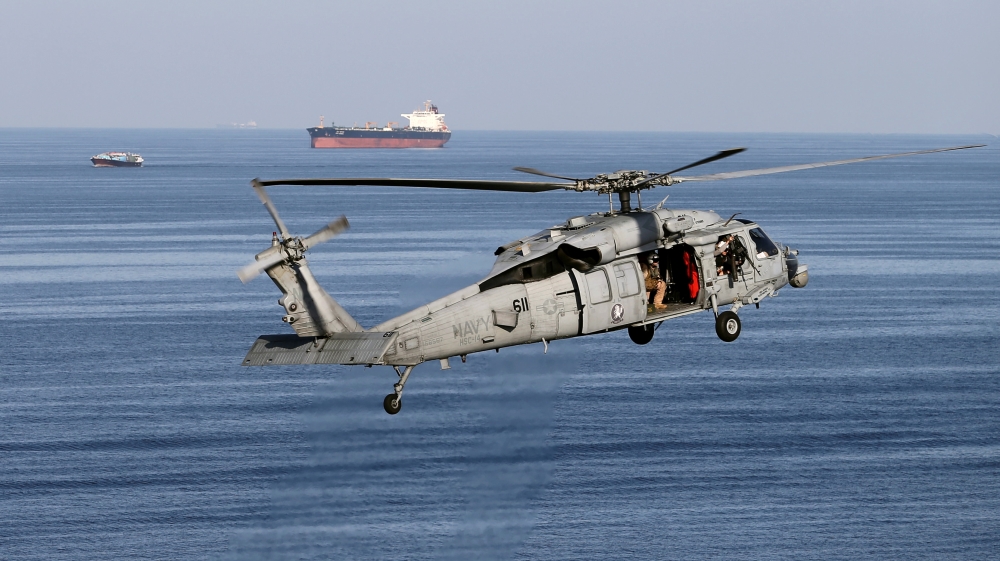MH-60S helicopter hovers in the air with an oil tanker in the background as the USS John C. Stennis makes its way to the Gulf through the Strait of Hormuz