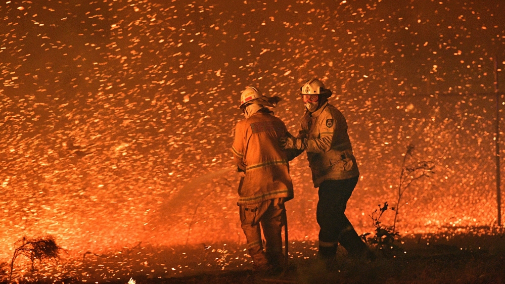 Firefighters struggling against the strong wind in an effort to secure nearby houses from bushfires near the town of Nowra in the Australian state of New South Wales. [Saeed Khan/AFP]