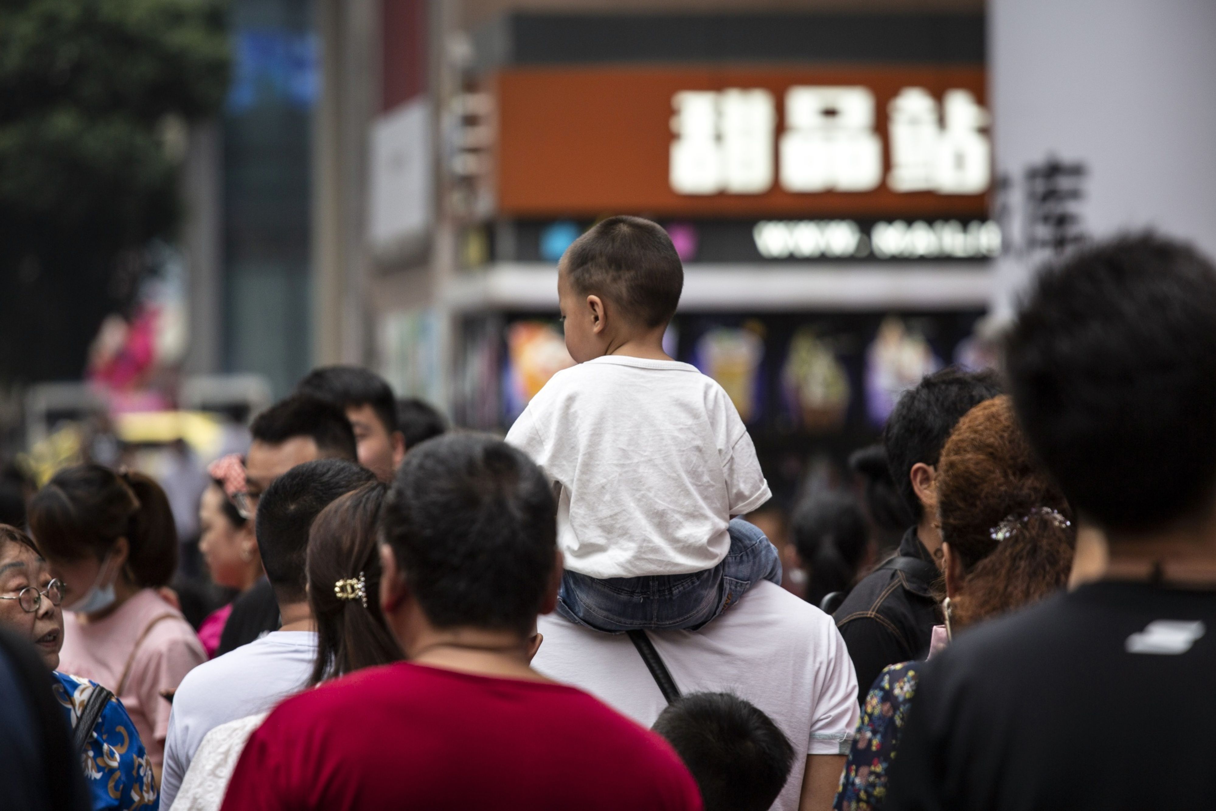 A child is carried on a man's shoulders in the middle of a crowded city sidewalk.
