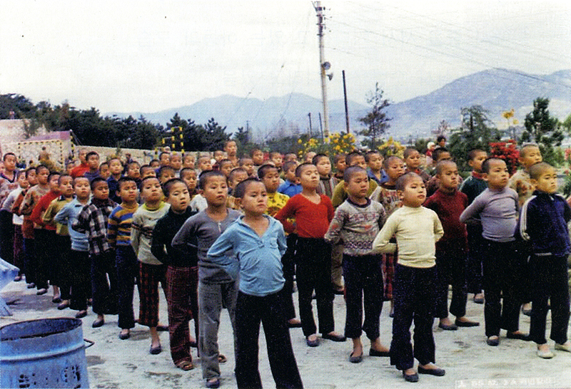 Children with shaven heads stand in queues, with hands behind their backs.
