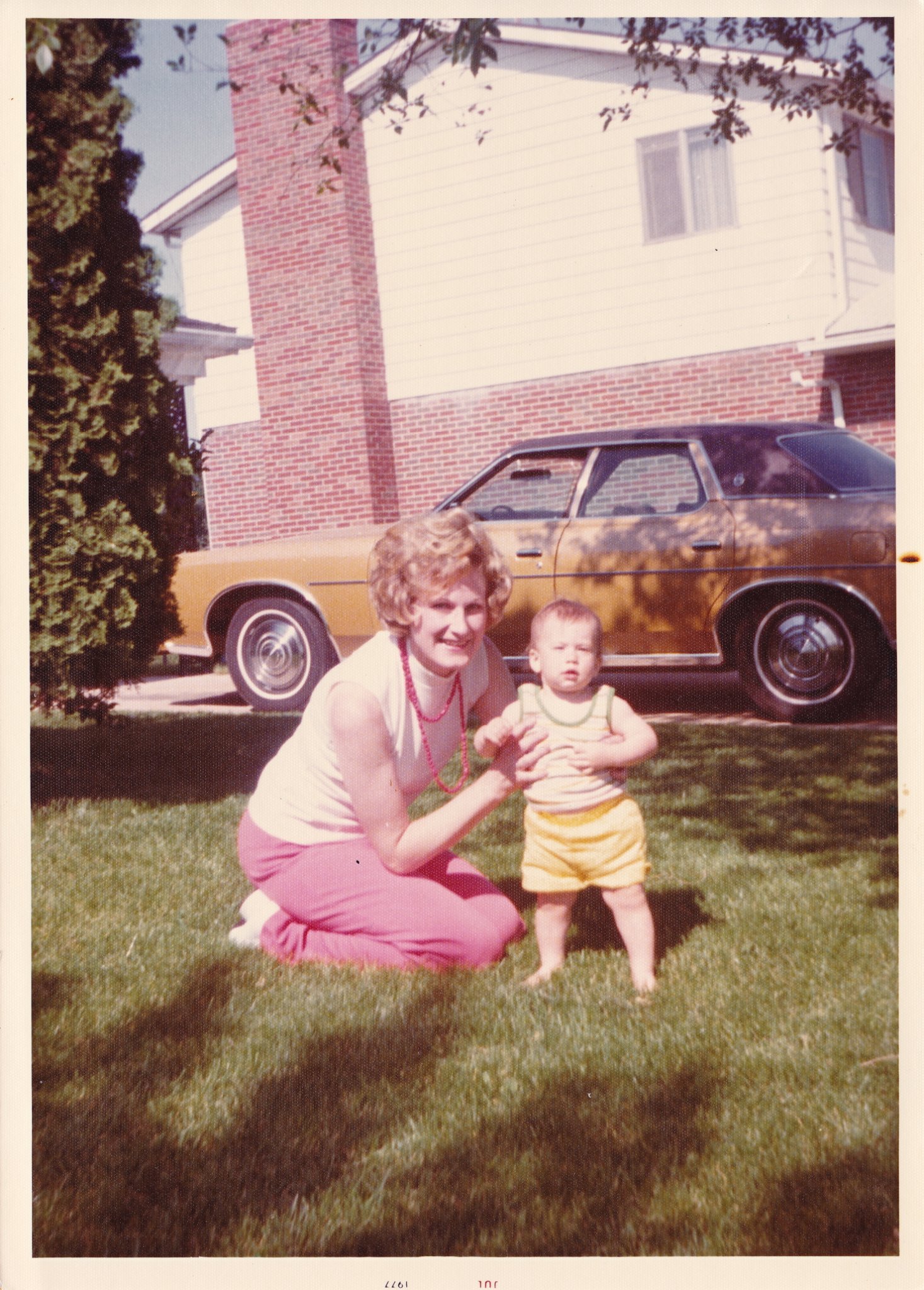 A photo of a woman sitting on grass while holding a baby that is standing up on the grass in a neighborhood.