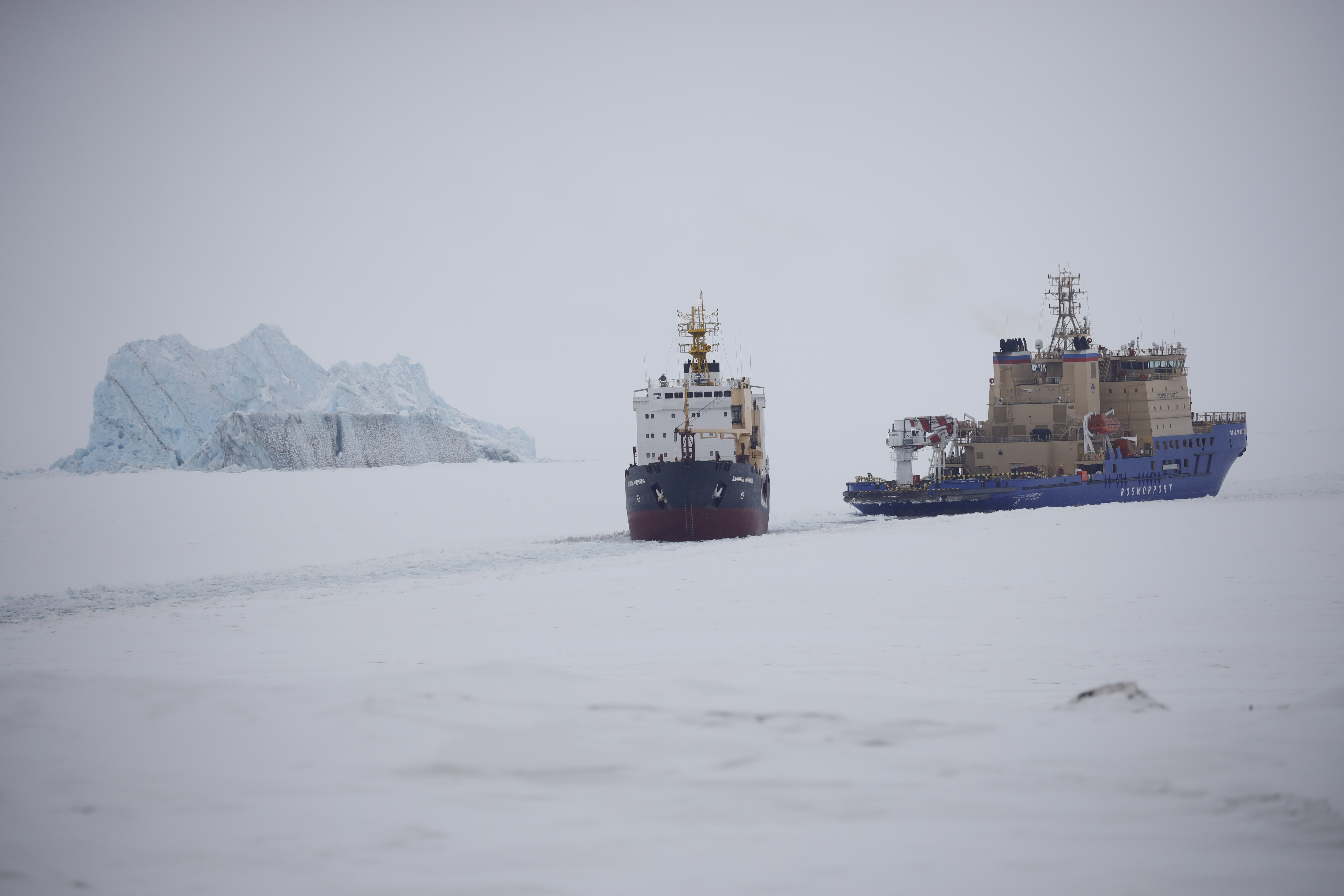 An Icebreaker making the path for a cargo ship with an iceberg in the background near a port on the Alexandra Land island near Nagurskoye, Russia, Monday, May 17, 2021.