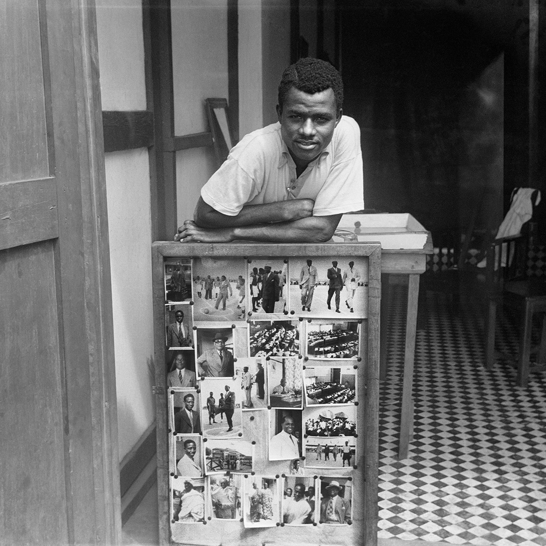 A photo of a man leaning against a table with a board in front of it with photos pinned to it covering the space.