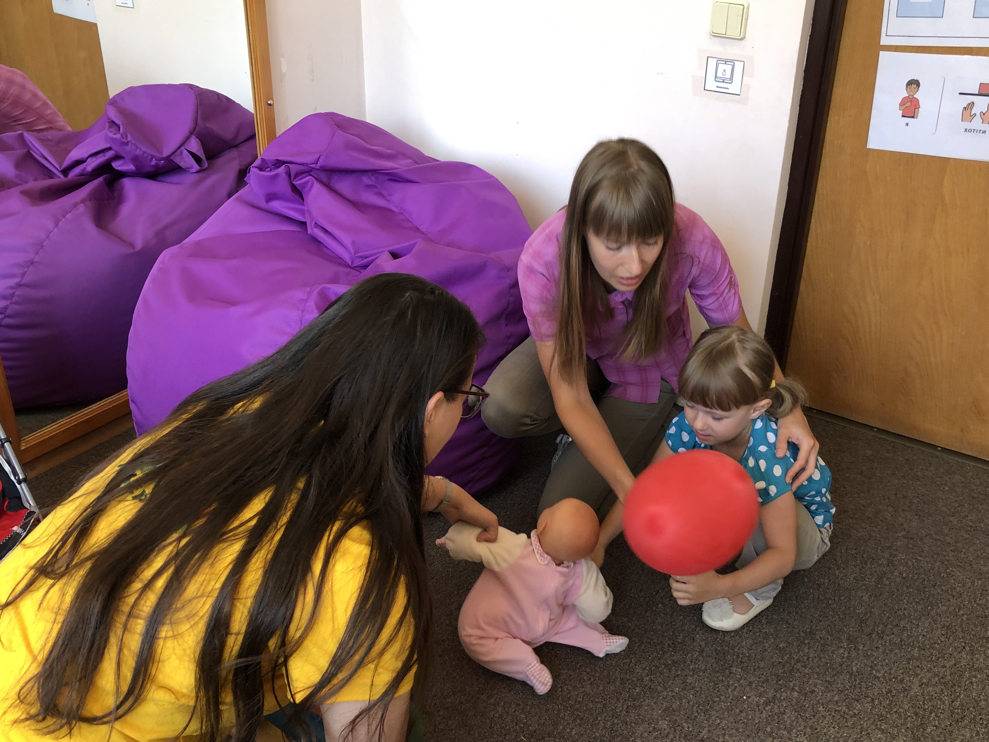 A photo of Viktoria Plyush holding her daughter while her daughter holds a balloon and reaches for a baby doll a woman in front of her is holding.