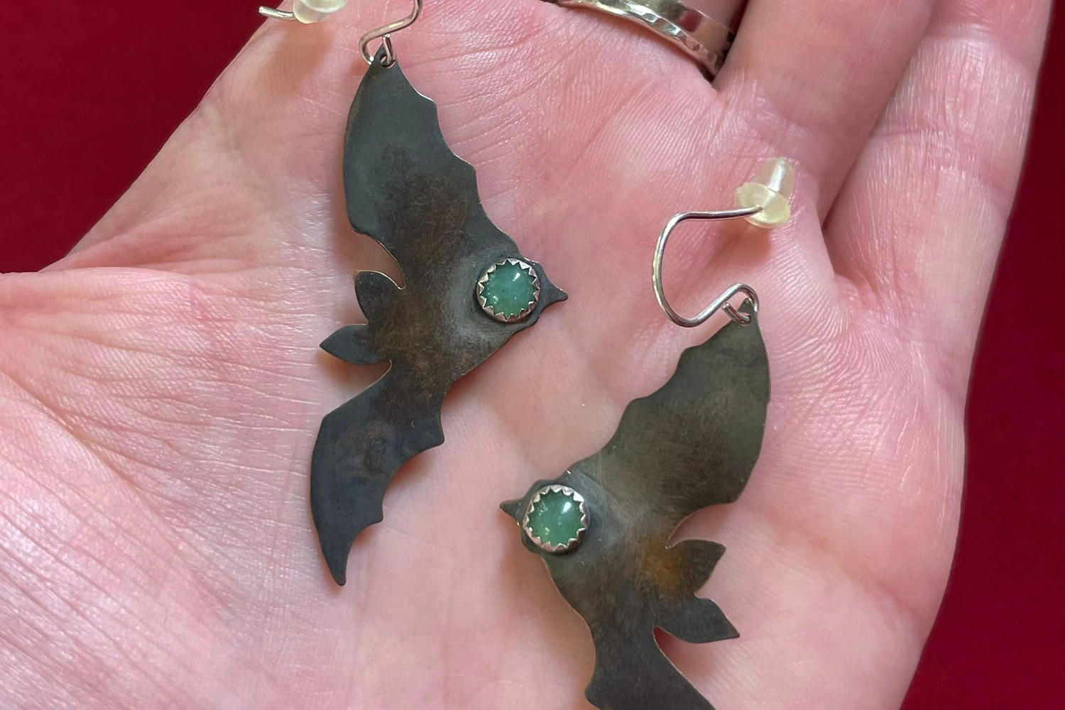 A photo of a person holding a pair of earrings with a bat shape cutout hanging off the earrings with a green stone on the end of the bat.