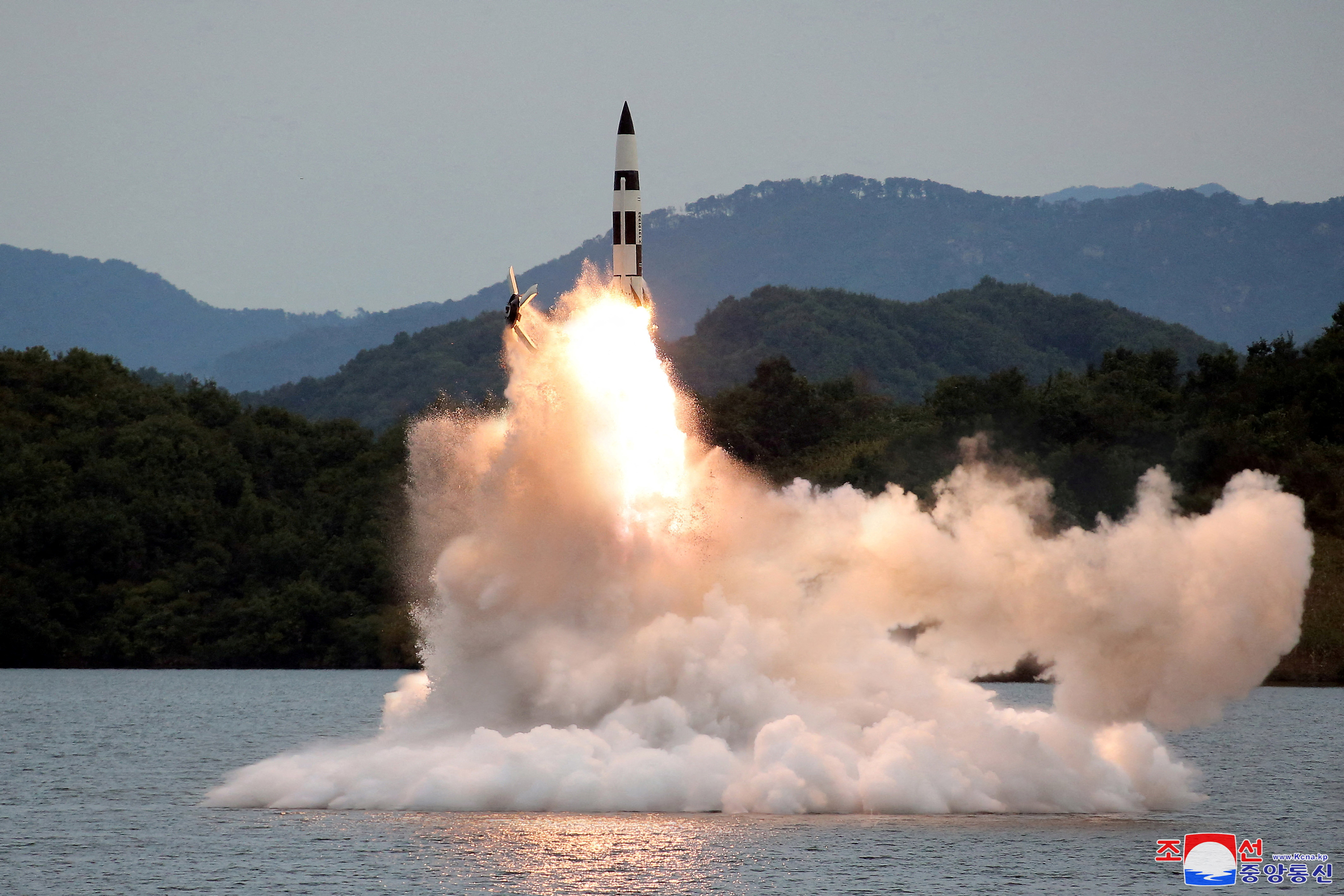 A missile launched into the air in clouds of white smoke from what appears to be a lake with mountains behind.