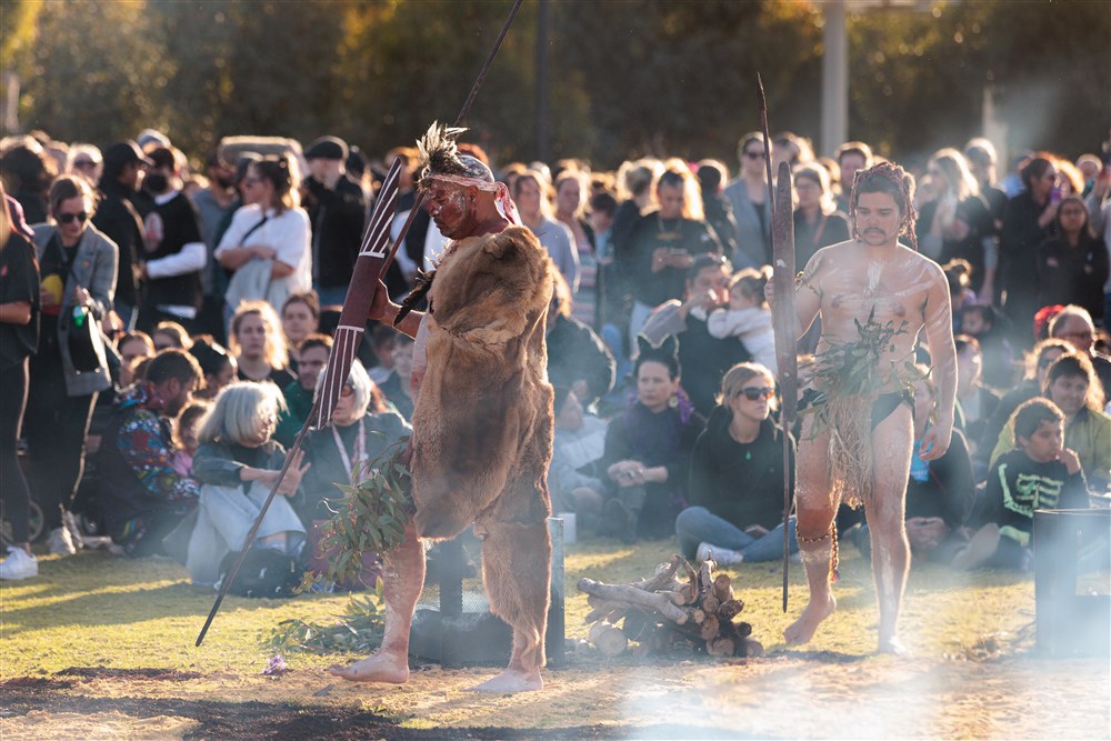 Two men in traditional dress walk past some wood on the ground. There is smoke all around them as they also carry large sticks and what look to be wooden shields. People stand and sit behind them watching the ceremony