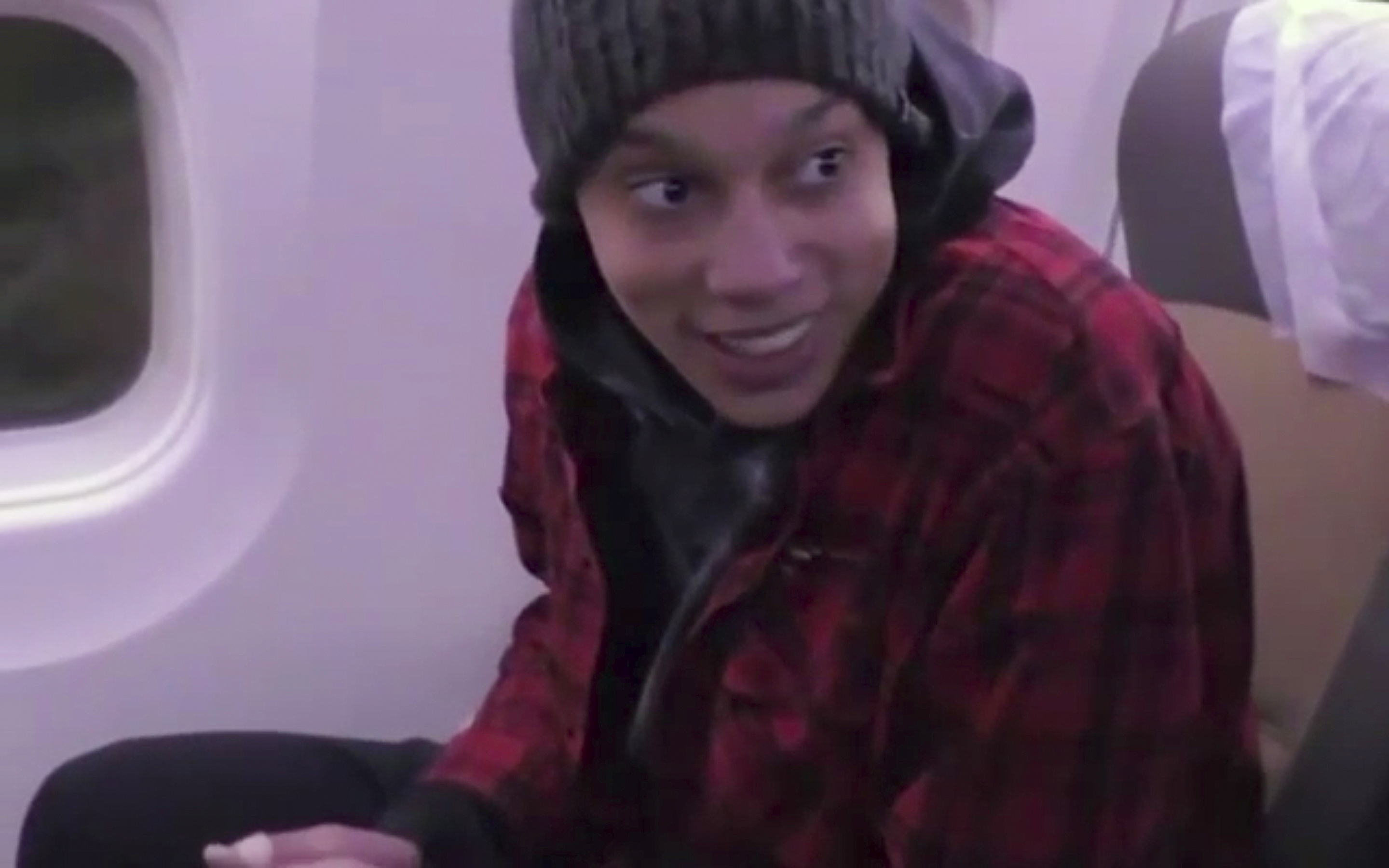 Brittney Griner smiling as she sits on a plane on her way out of Russia. She's wearing a red jacket and grey beanie