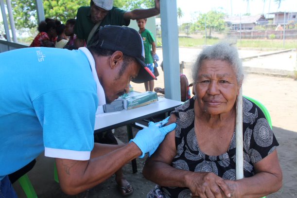 Woman with grey hair sits on chair, looking at the camera while a man bends over to administer the vaccine. He is wearing a cap, blue t-shirt and blue gloves. There are some people waiting behind them, including children