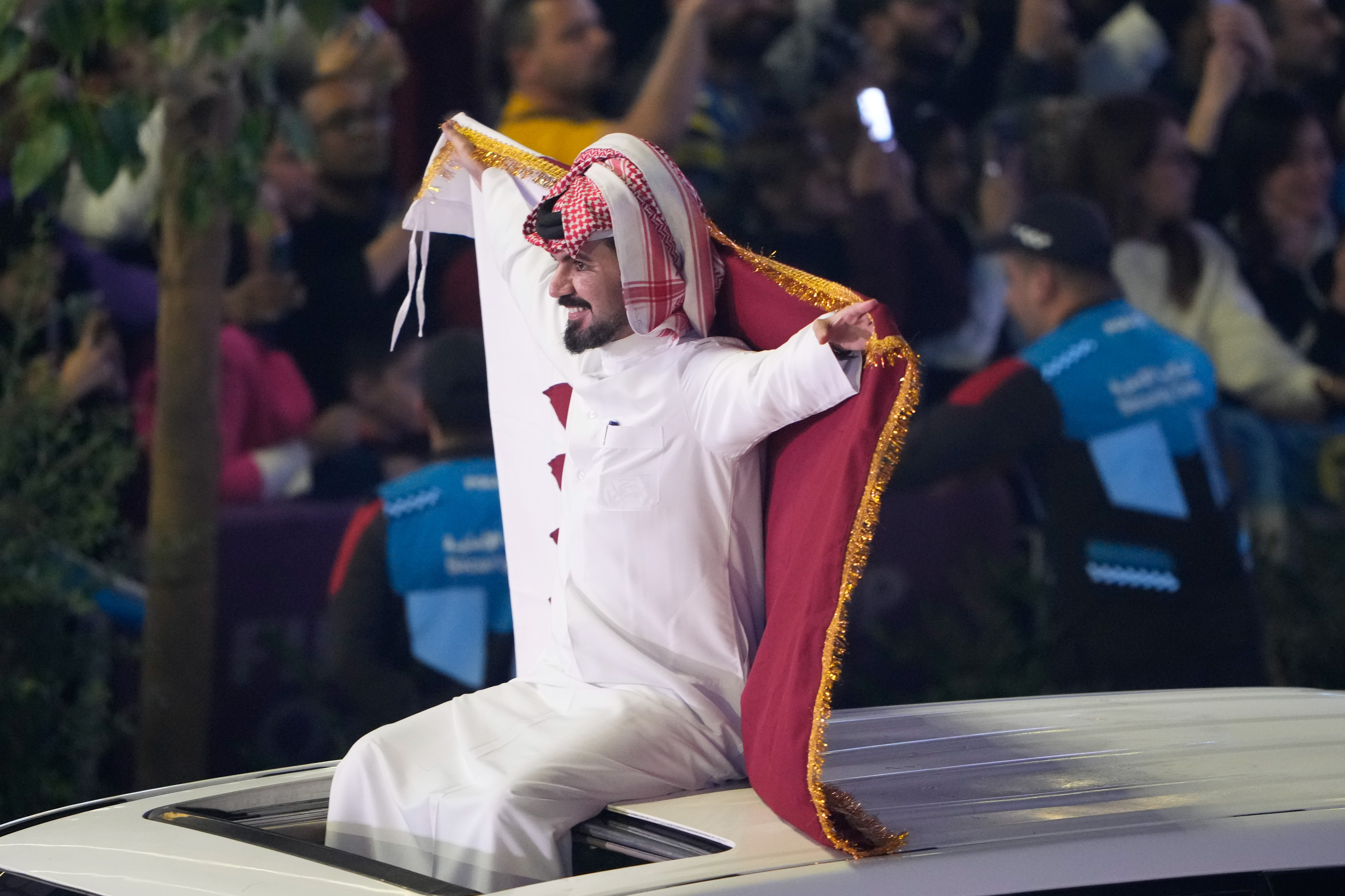 A man holds a Qatari flag while riding out of a vehicle's sunroof.