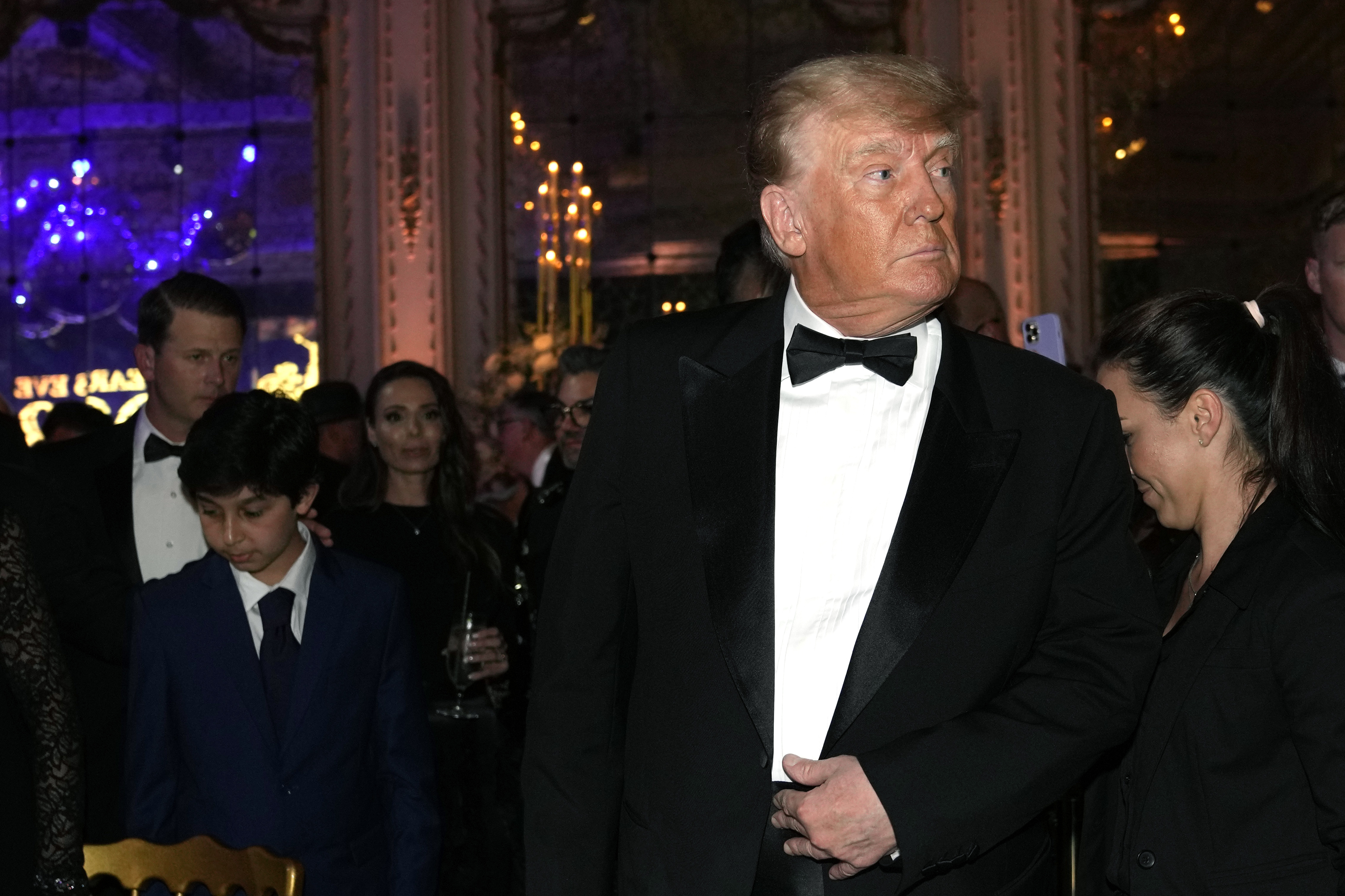 Trump wearing a tuxedo looks left. There are people behind him, one holding a champagne glass
