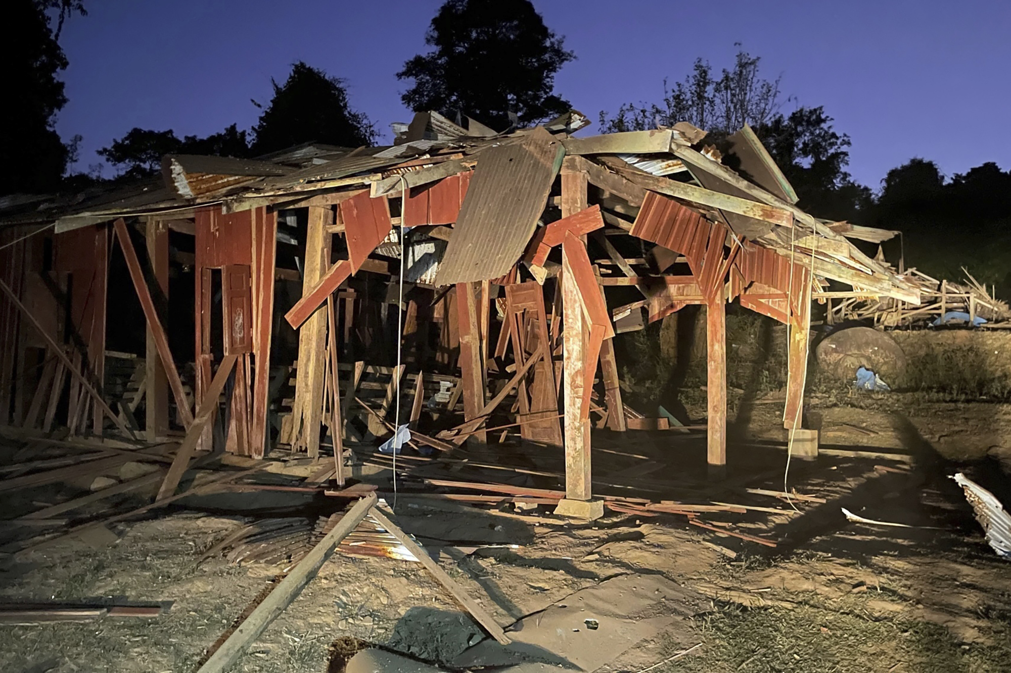 The ruins of a village school. Some wooden boards are still standing, but the wooden structure has generally collapsed. The sky is a deep purpose. It looks to be nighttime.