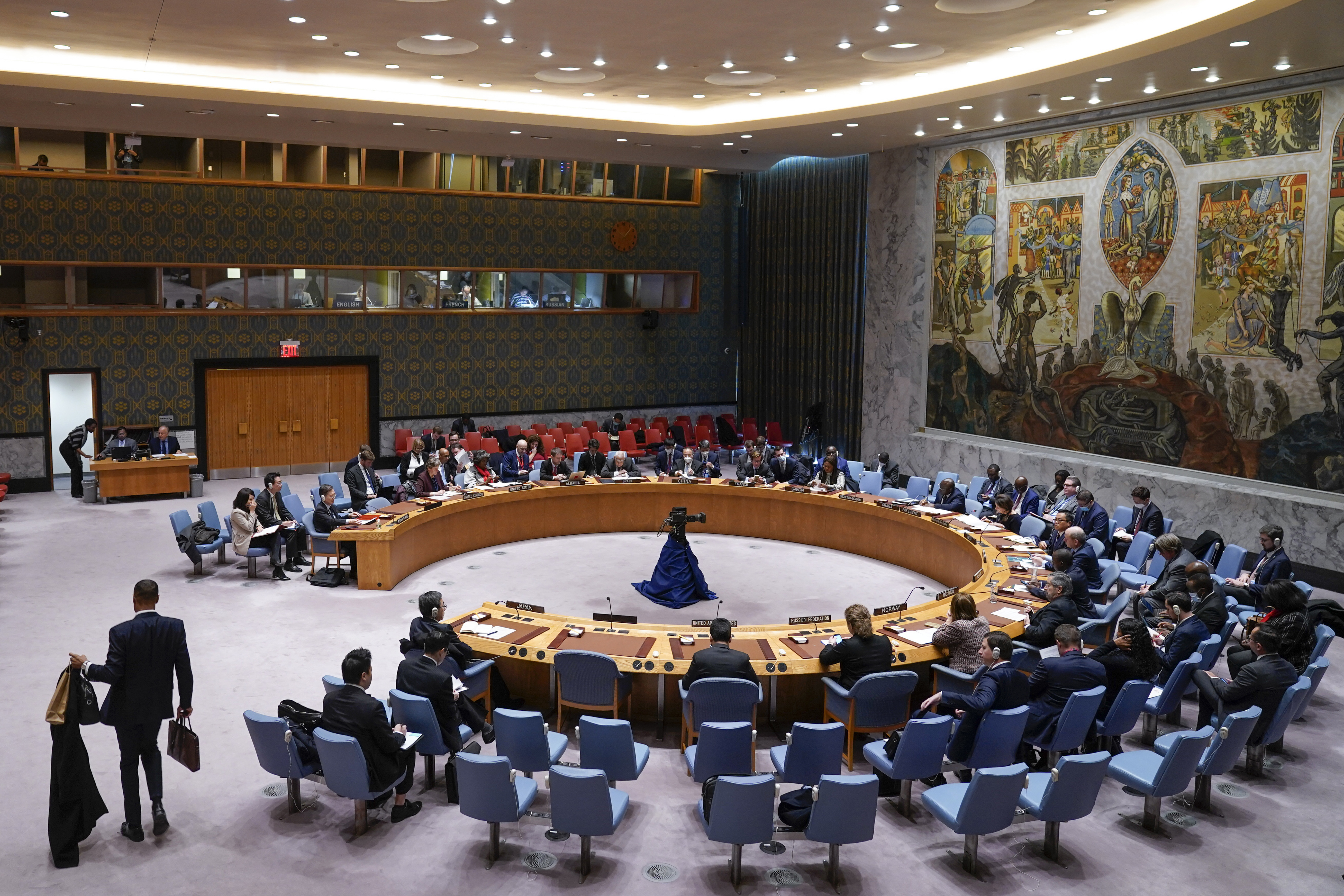 A wide view of the UN Security Council chamber. There are delegations around a central horseshoe shaped table and a large mural on the wall behind the table.