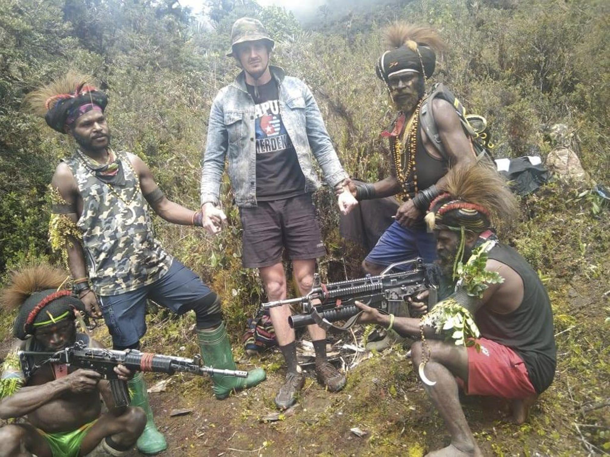 A man said to be Phillip Mehrtens surrounded by rebels in a rural location. He is wearing a t-shirt for Papuan independence, denim jacket, brown shorts and a hat. One rebel fighter is holding his wrist and another his hand. There are two fighters squatting at the front holding their guns.