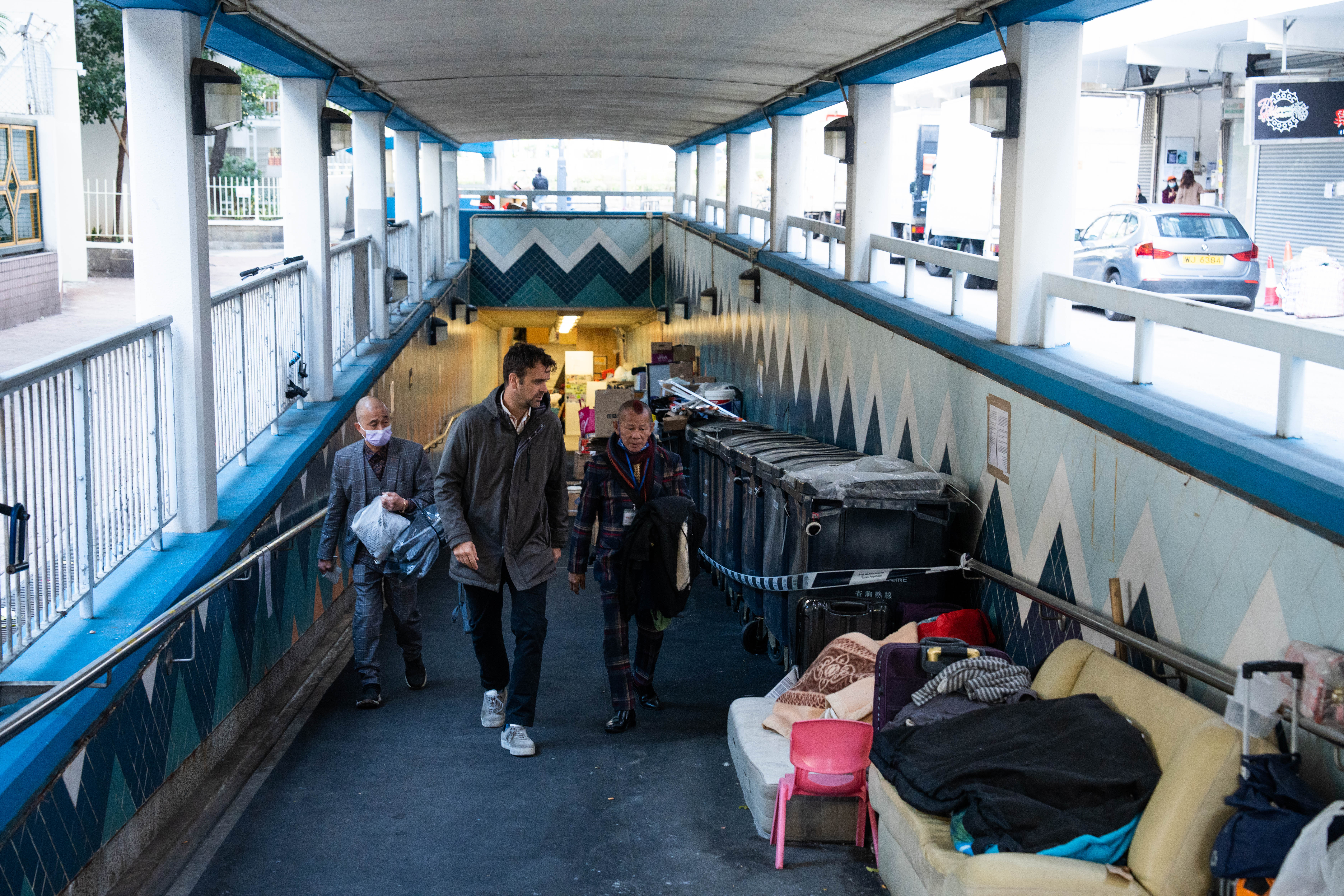 ImpactHK founder Jeffrey Rotmeyer and one of his staff walking out of an underpass where many homeless people live. There are old sofas and furniture that people have saved from rubbish dumps. There are cars on the road outside.