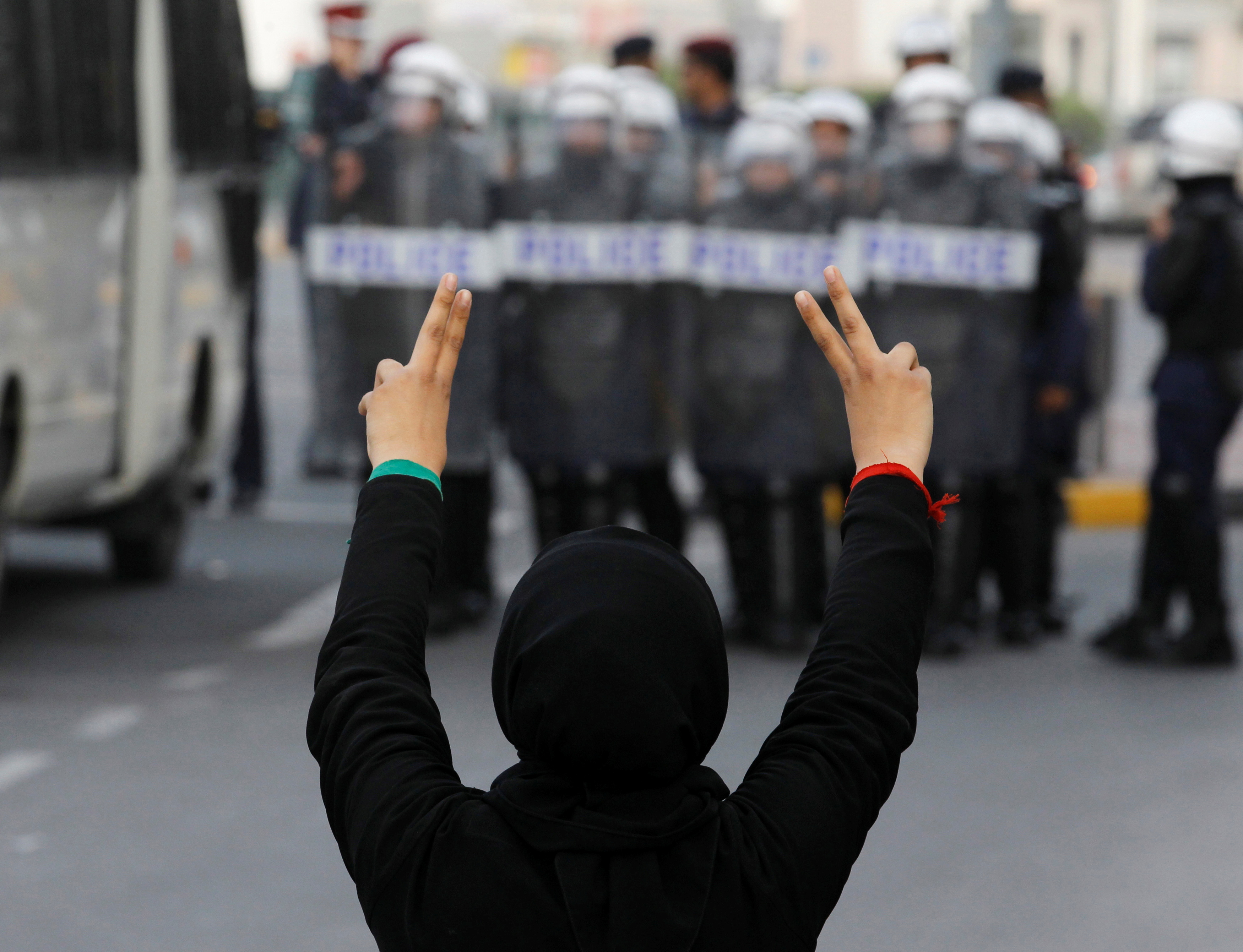 A woman holding up her fingers in a V for Victory sign towards a row of riot police in Manama, Bahrain. She has her back to the camera, is wearing black and has red and green ties around each wrist