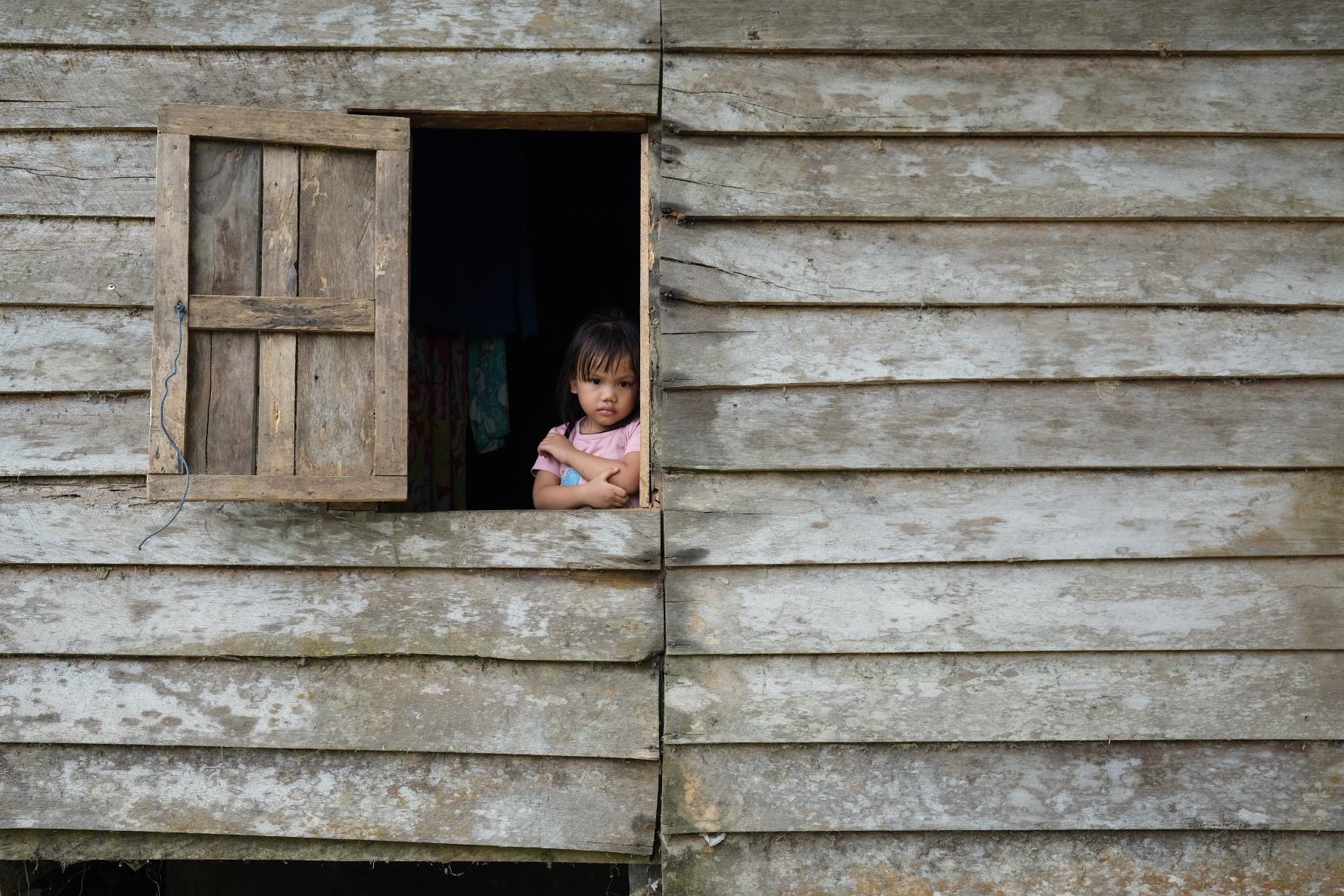 A young girl looks from the window of a wooden house. She is wearing a pink t-shirt and has her arms around her chest. She looks quite serious.