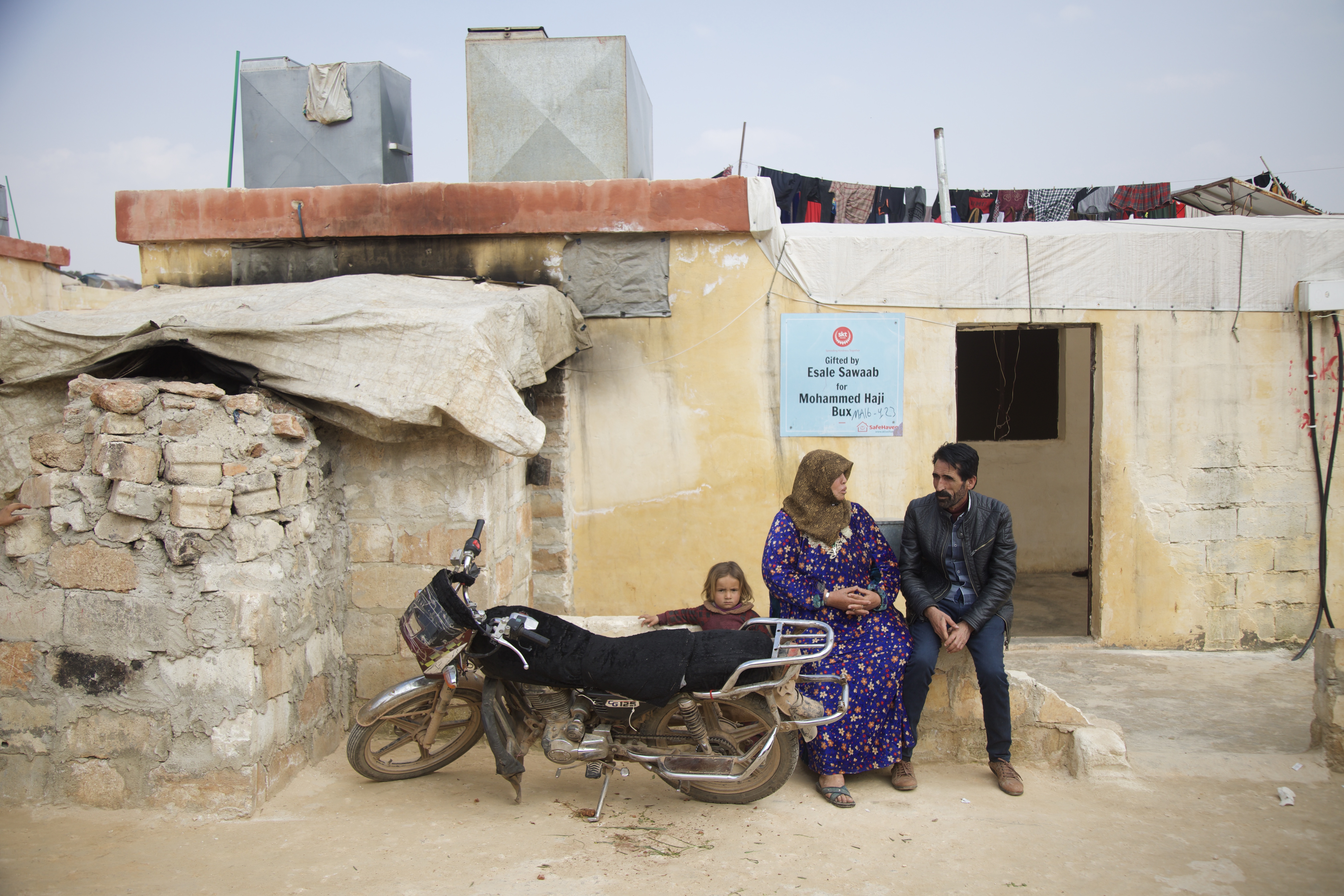 A man and a woman talk while sitting in front of a house, with a motorcycle parked to their side.
