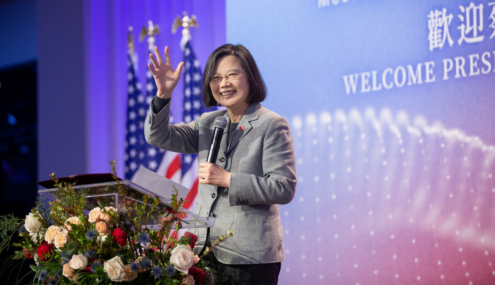 Taiwan President Tsai Ing-wen waves during a meeting in New York with the Taiwanese community. She is on a stage behind a lectern and smiling. There is a large sign at the back of the stage welcoming her.