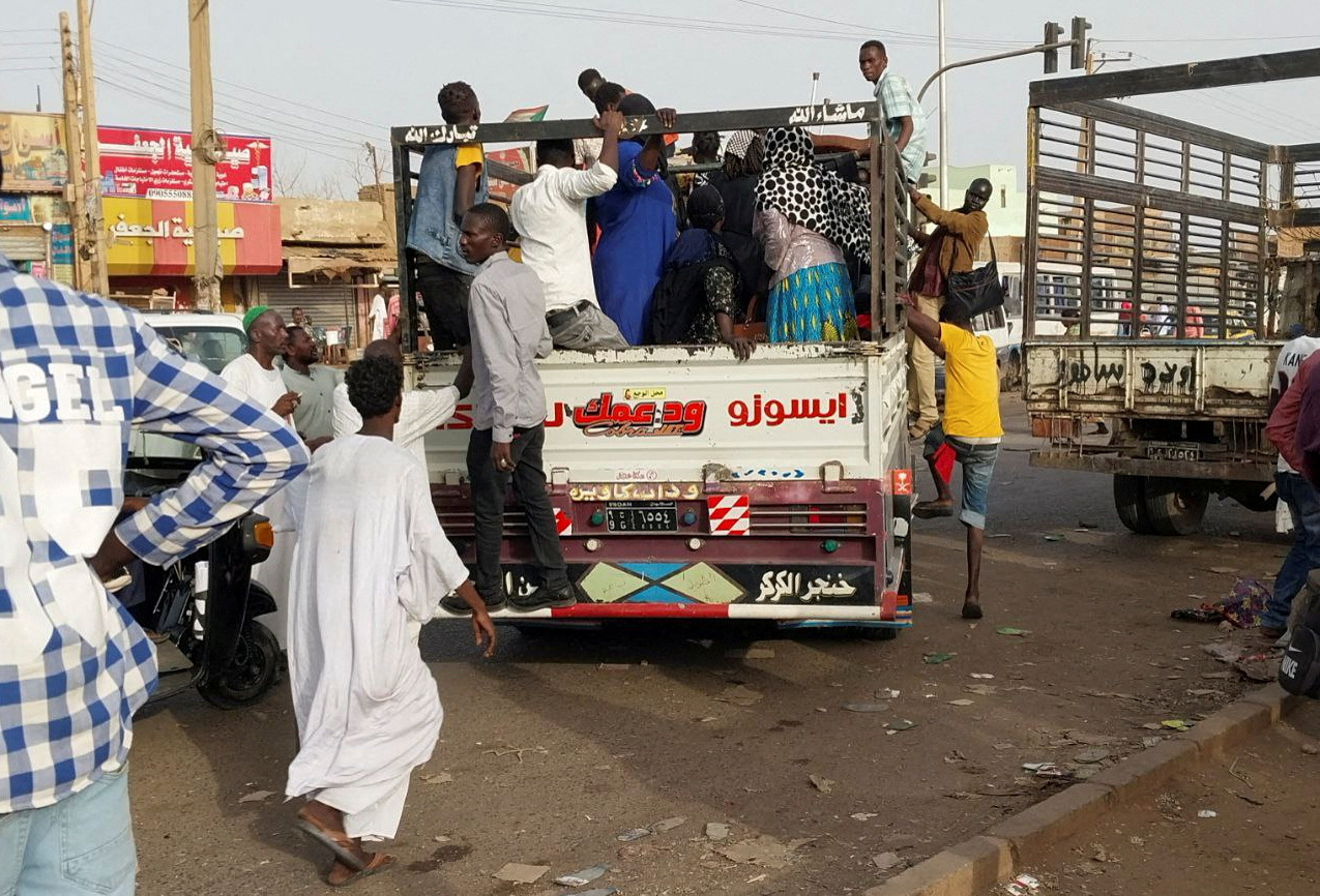 People getting onto trucks to try and leave northern Khartoum. Some are hanging on the side of the vehicle. One man is running to get on board as the truck begins to move.