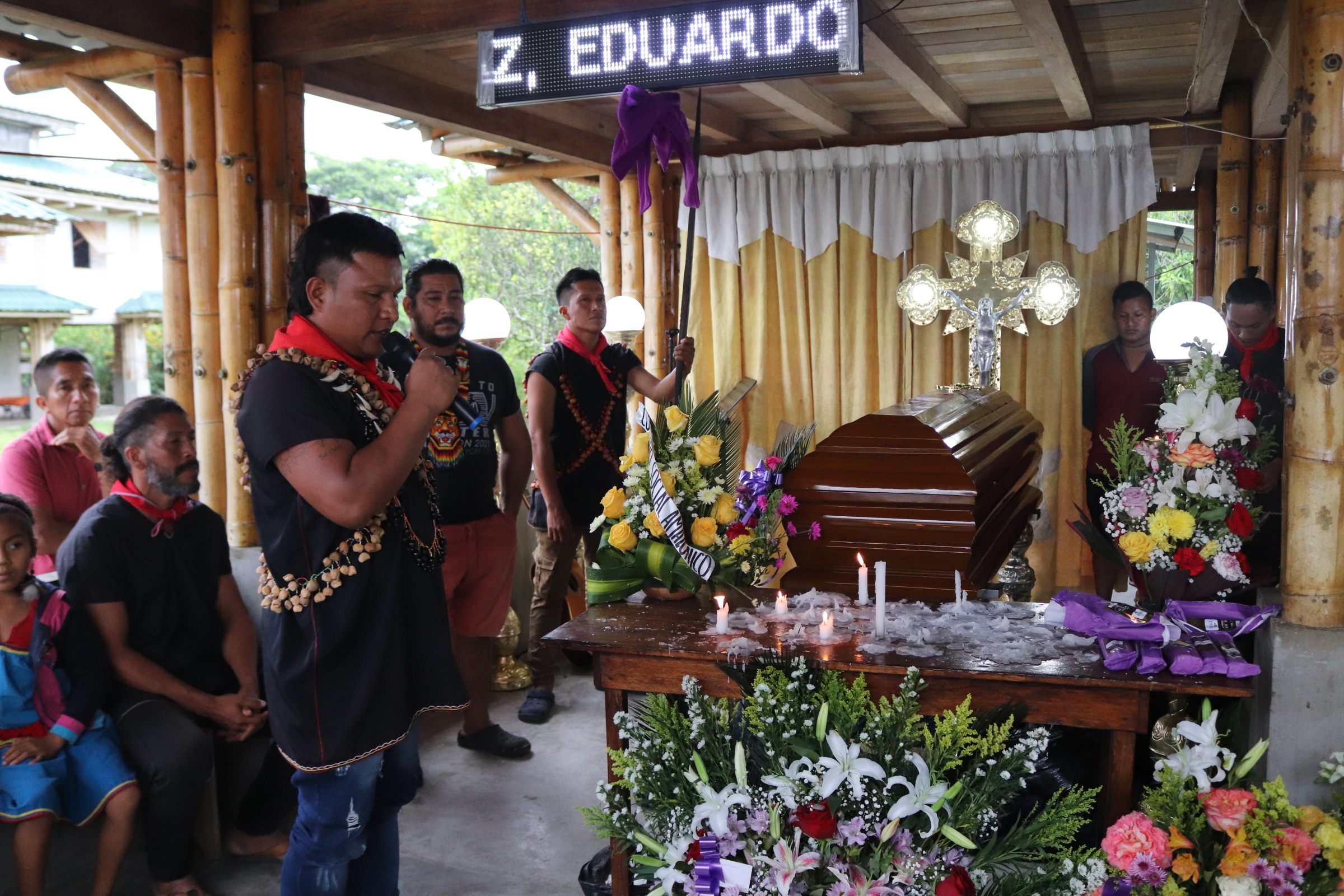 A person stands praying next to a coffin, decorated with flowers before a cross