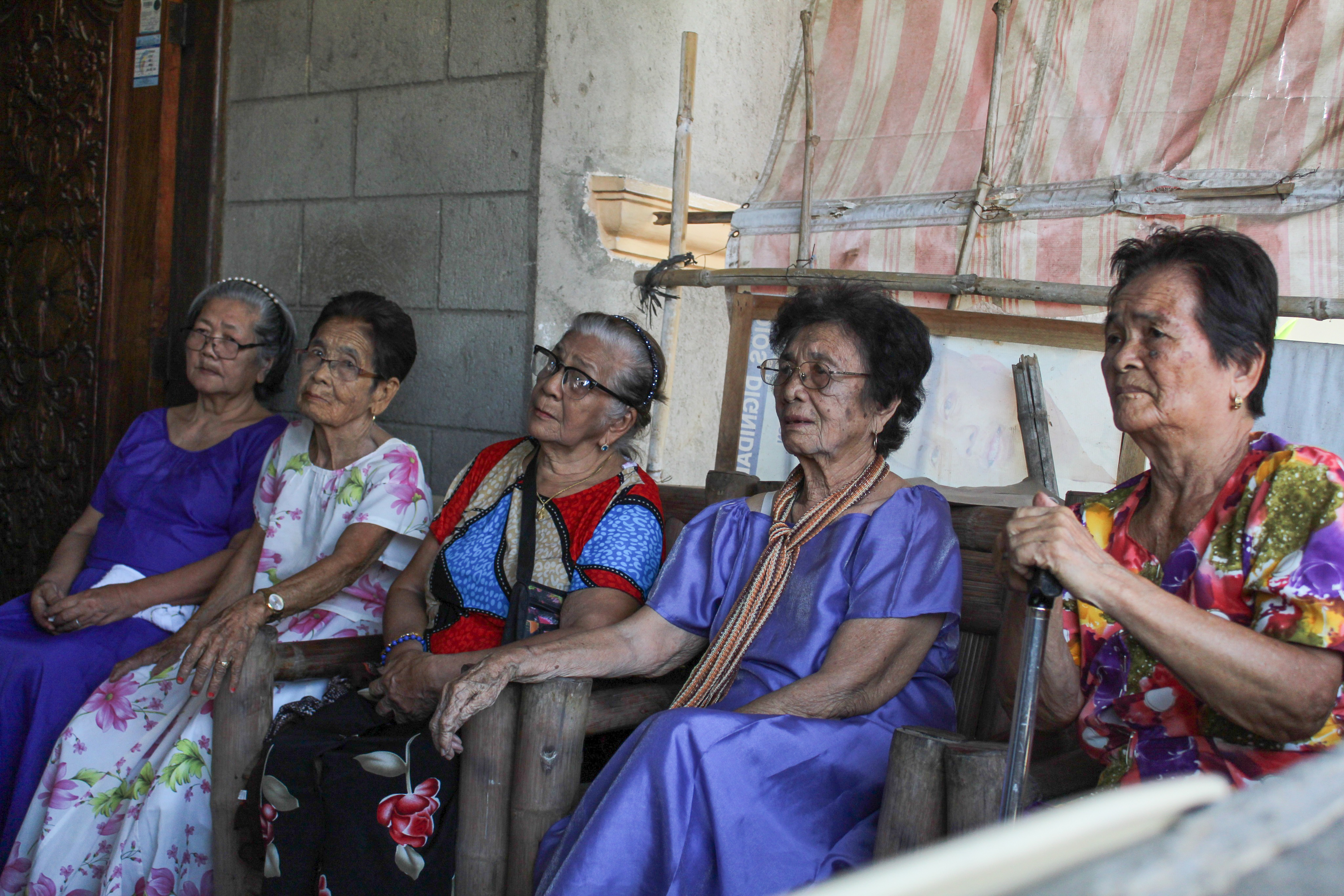 The women from the Malaya Lolas. They are sitting together in a row on a chairs made from bamboo. They look thoughtful. One woman has her hands on a walking stick and looks upset. 