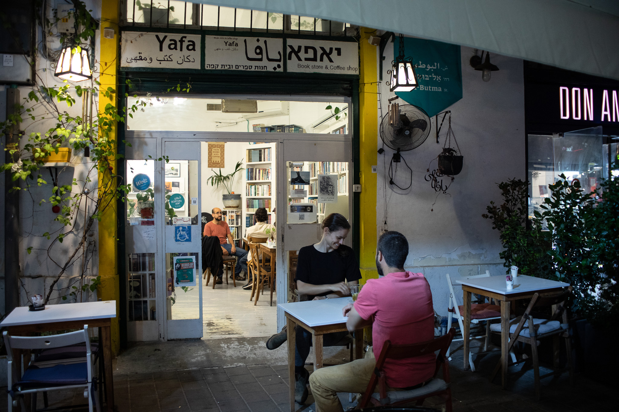 two people sitting and chatting at a table outside, while the open door shows more people talking inside in front of packed booskelves