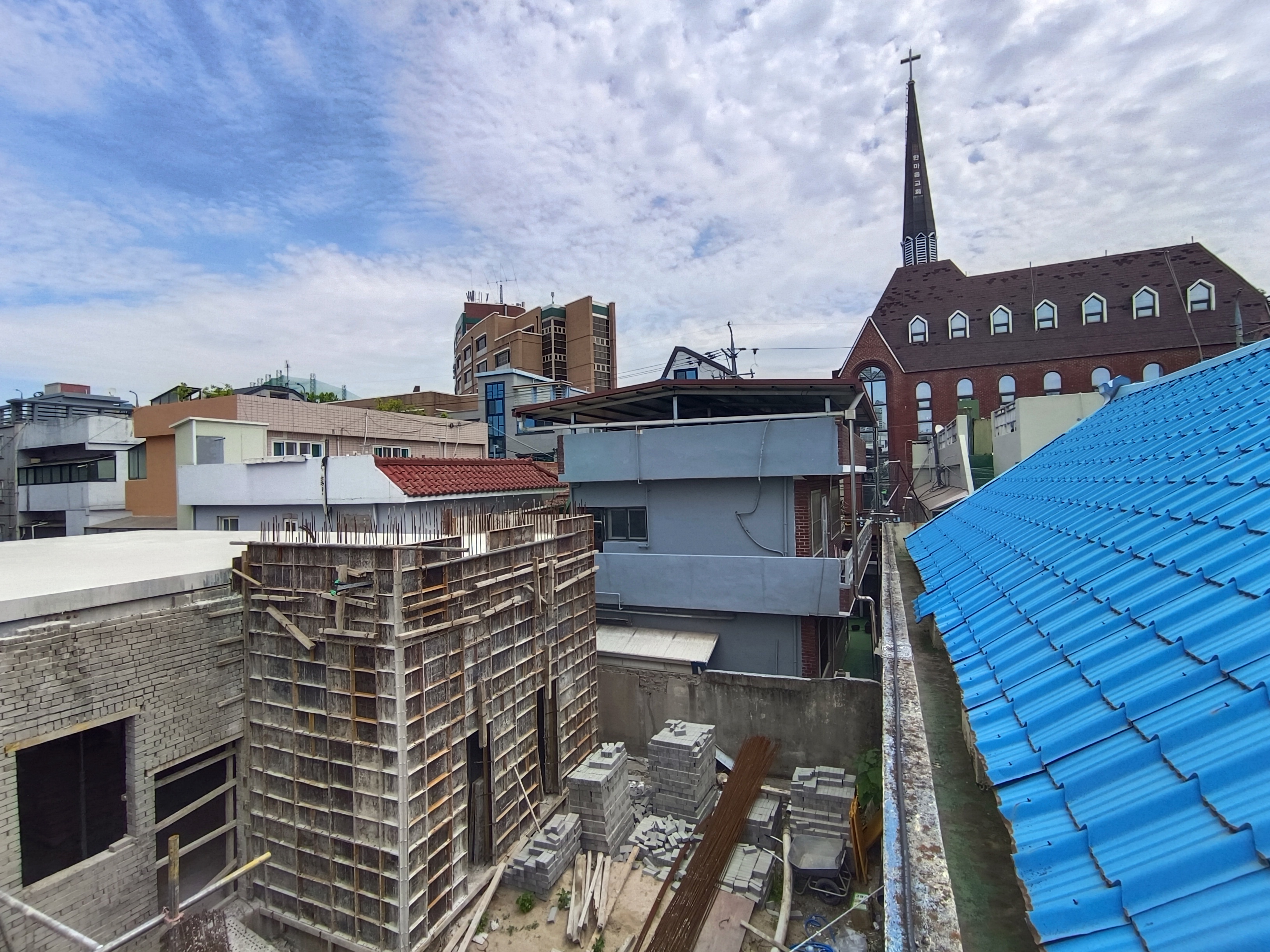 View of roof tops from the temporary prayer room at the mosque showing the construction site. There is a church with slender spire and a cross on top 30 metres behind, and lots of tightly packed low-rise buildings.