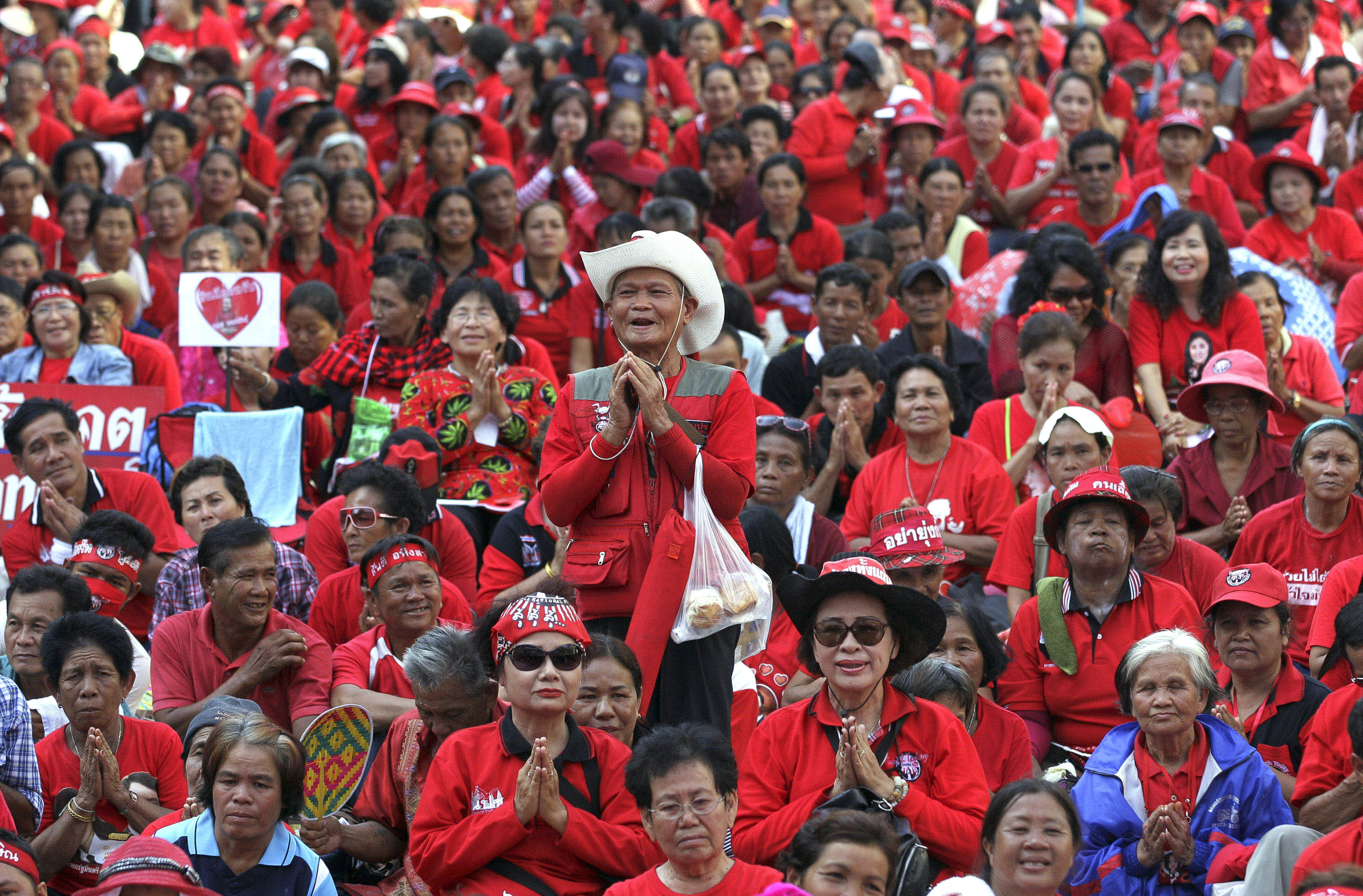 A group of protesters wearing red seated on the ground. There is a man standing in the middle of the photo. He is wearing a white cowboy hat and has his palms together in prayer. The crowd is also praying.