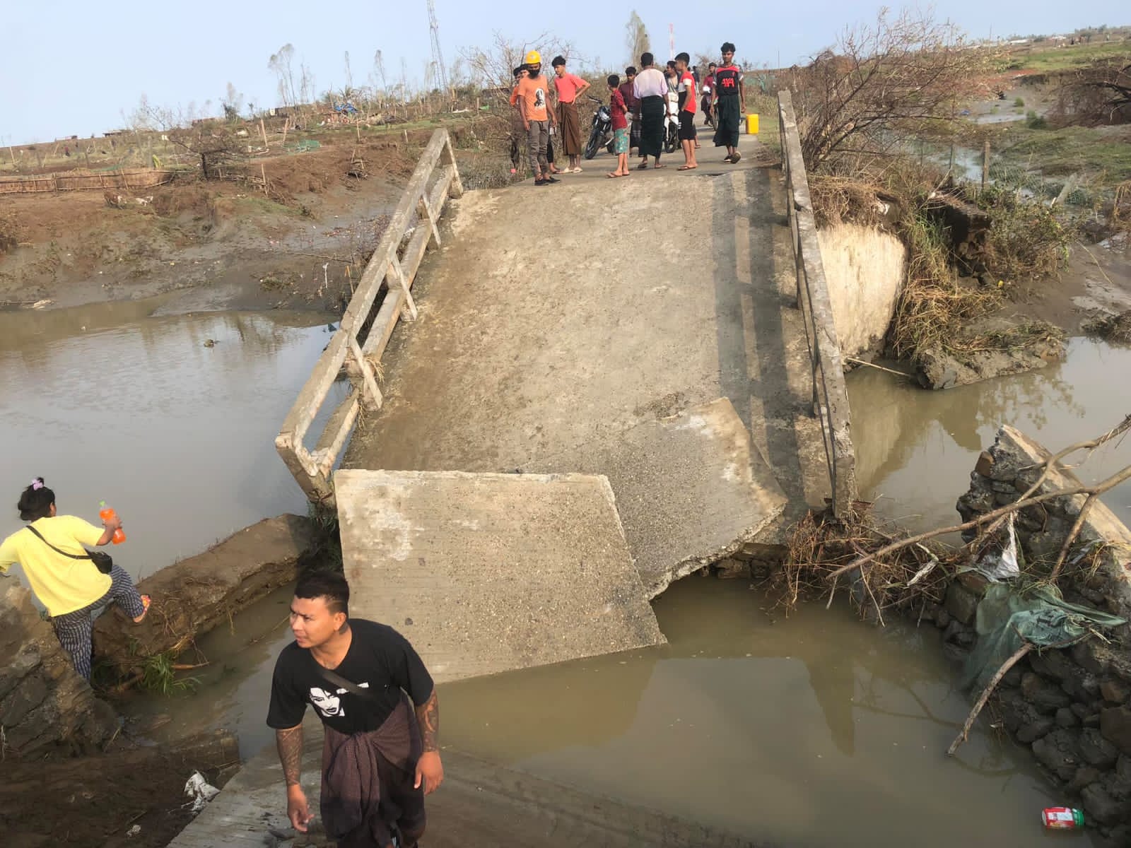 A concrete bridge broken by Cyclone Mocha between Sittwe and Kyauktaw. One man is standing in the water in front. Others are on the bridge behind. The land around is brown and looks battered
