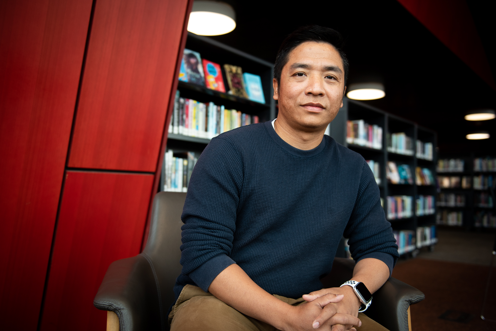 Chin community member Rual Thang. He is seated and wearing a black long sleeved T-shirt. There are shelves of books behind him.