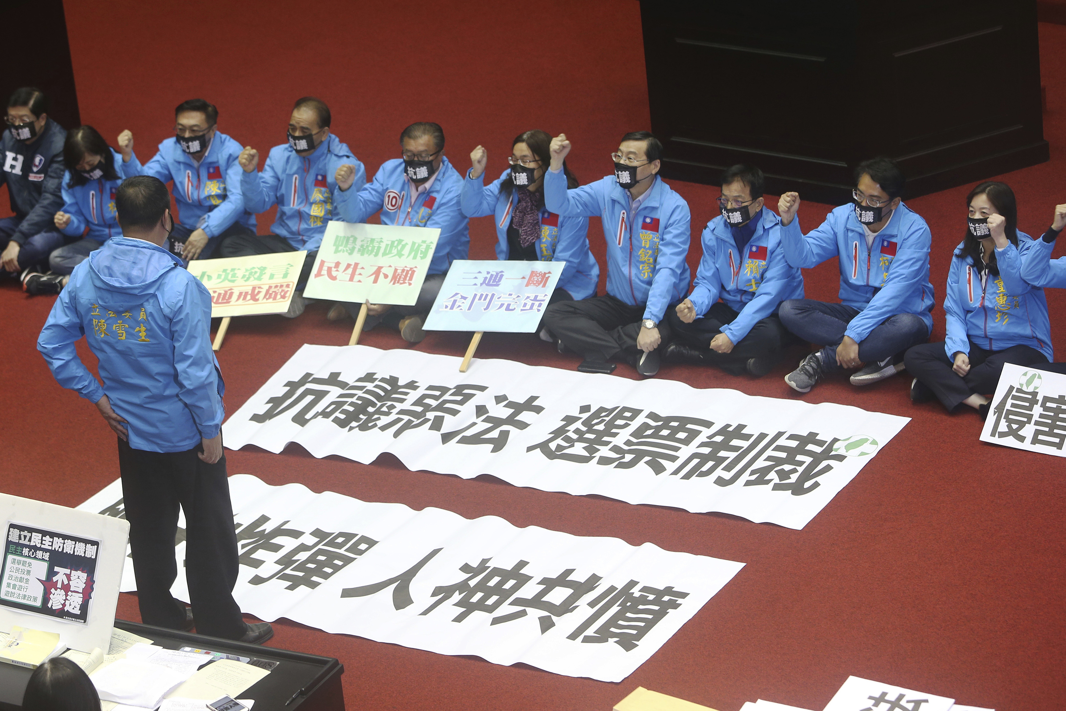 KMYT legislators dressed in sky blue bomber jackets sitting on the floor of the legislature in protest against the anti-infiltration bill. They have banners in Chinese characters reading 'Protest against a bad law", "Sanction by Votes. "Neck Bomb" "Be hated by both man and God''. The banners are laid out on the floor. Three are holding a banner in their hands.