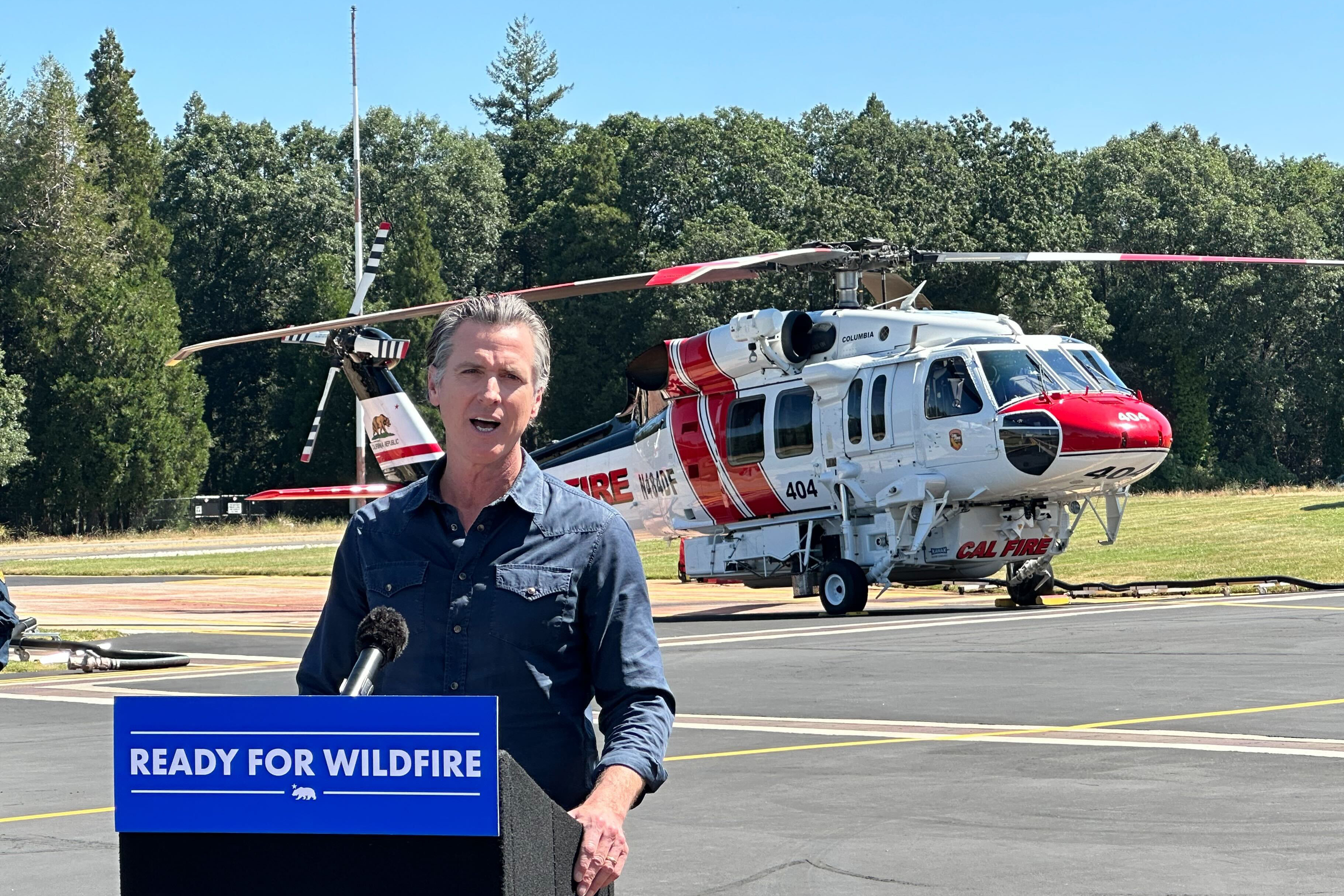 A man at a podium labelled "ready for wildfire" speaks and stands in front of an emergency helicopter