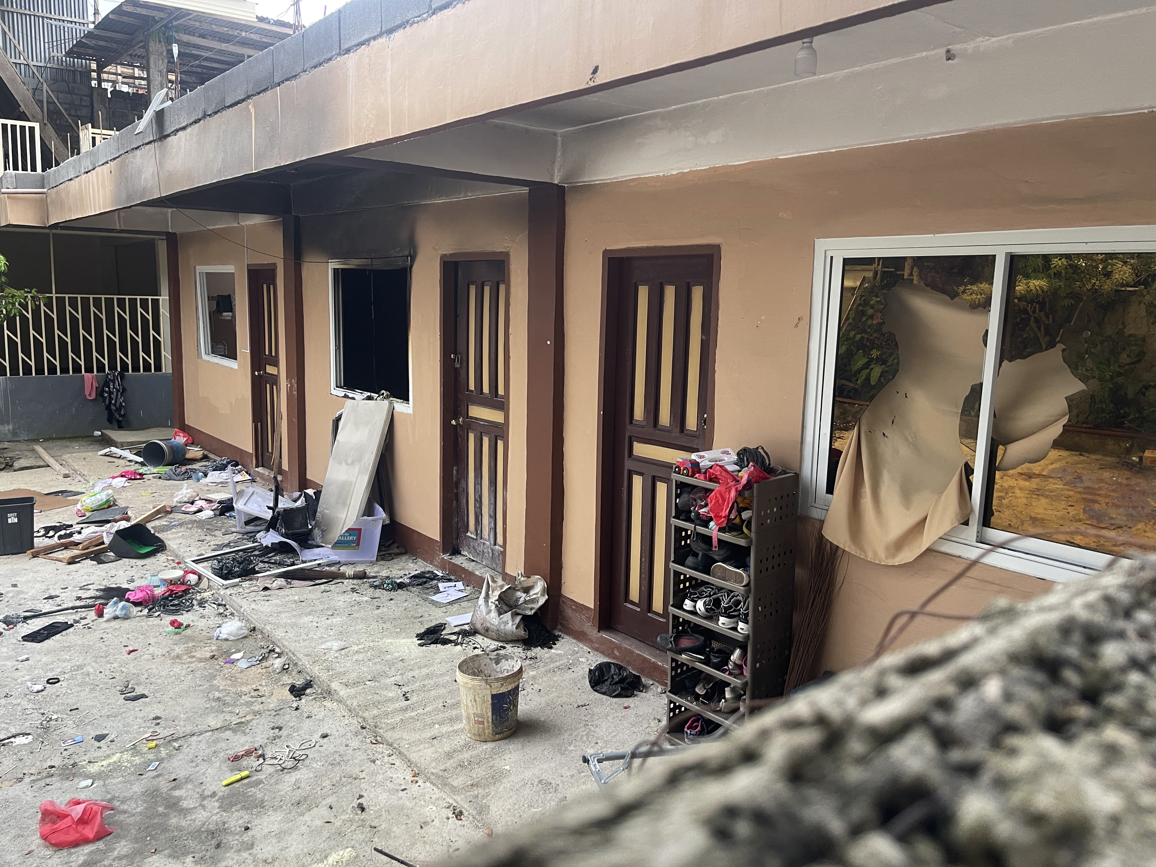 The exterior of the aoartment. The walls are painted a peach colour. There is shoe rack outside one of the doors. A window is missing and seems to have been blown out. There is bucket and other paraphernalia lying about