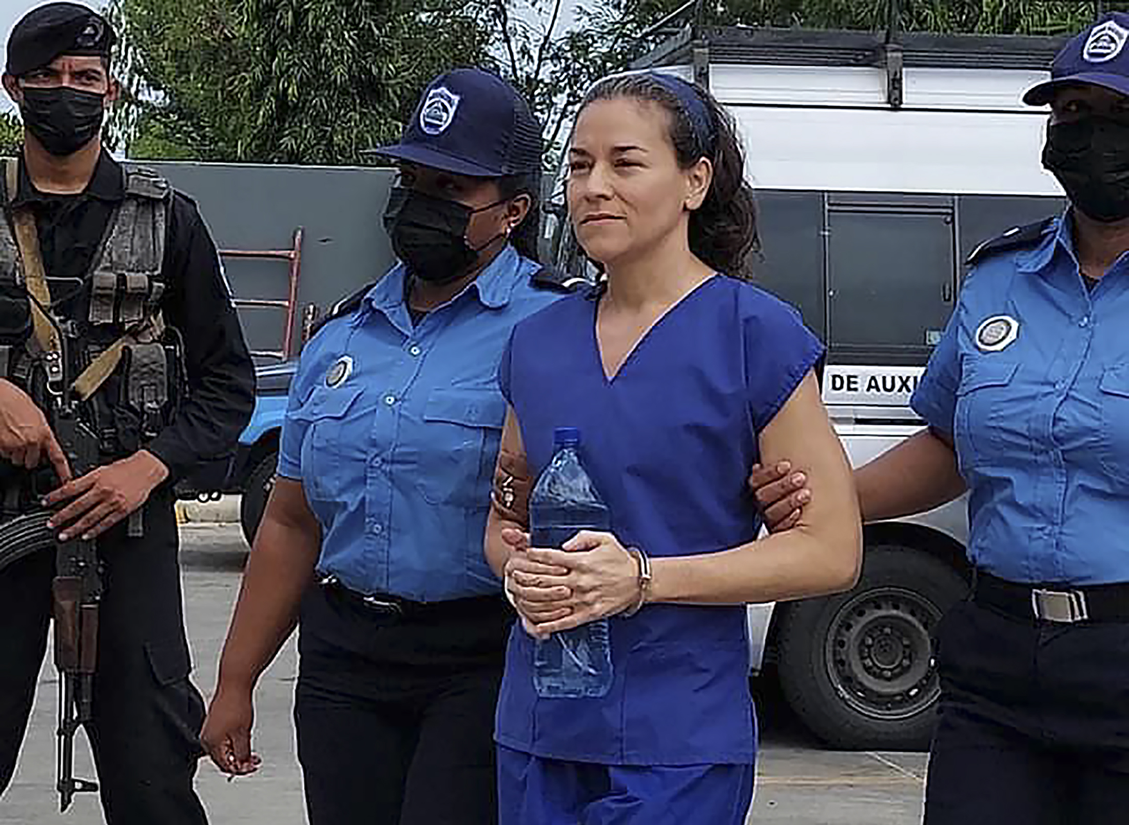 A woman in a blue prison jumpsuit arrives handcuffed with a police escort outside a courthouse. A police van can be seen behind her.