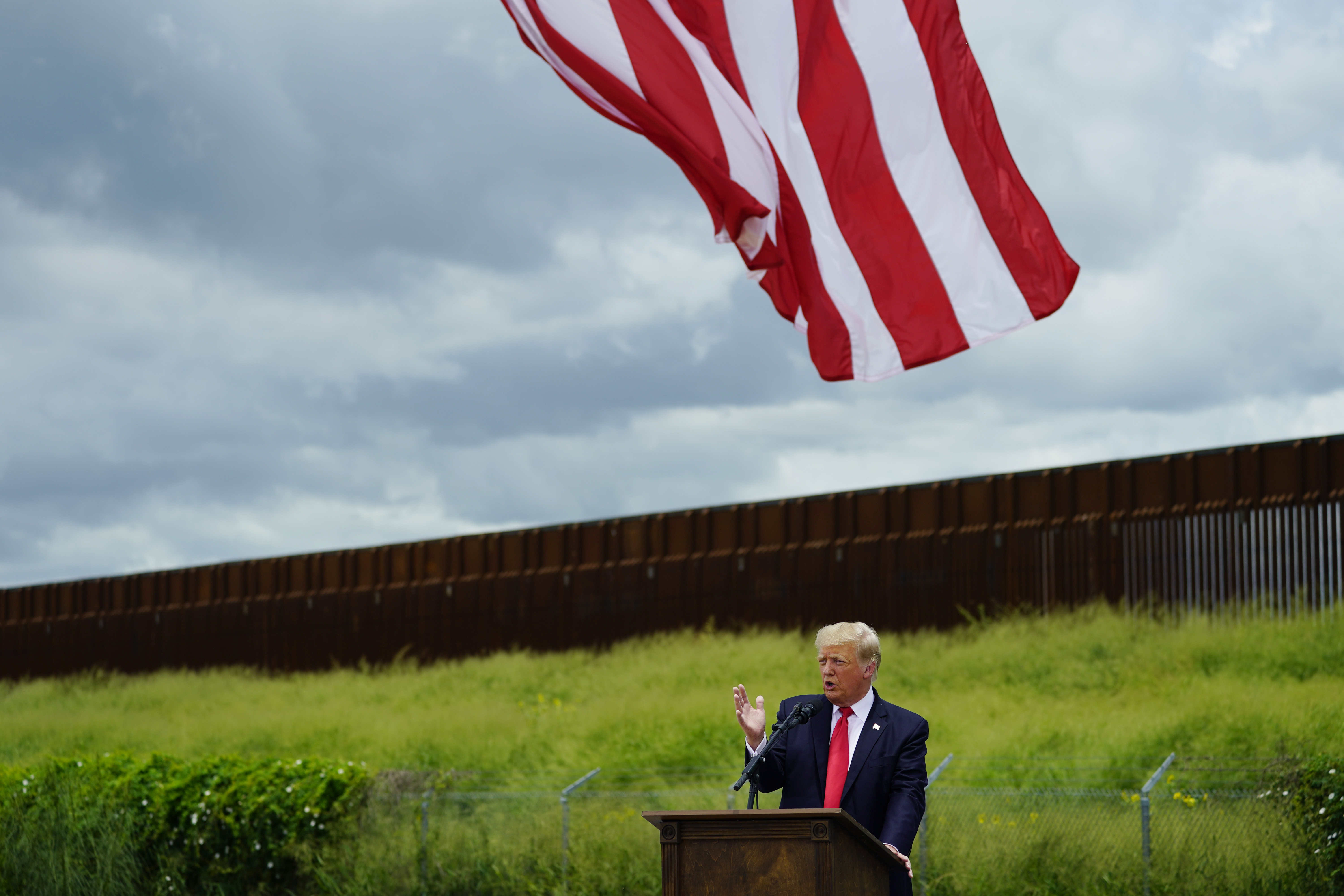 Donald Trump stands at a podium, speaking, as a US flag waves overhead and a grassy hill stands behind him, capped by a metal border barrier.
