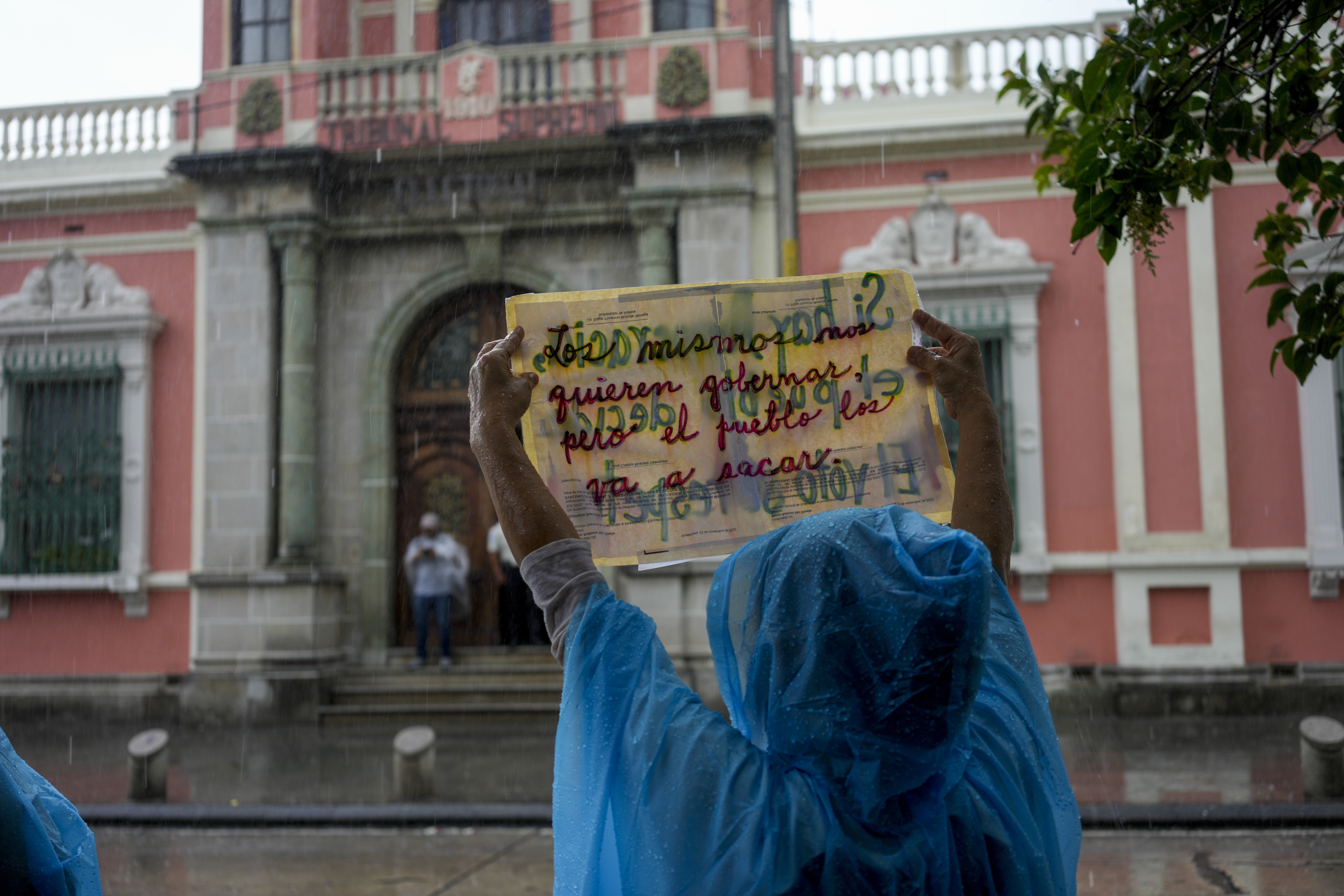 A woman in a blue rain poncho holds up a sign in front of a government building.