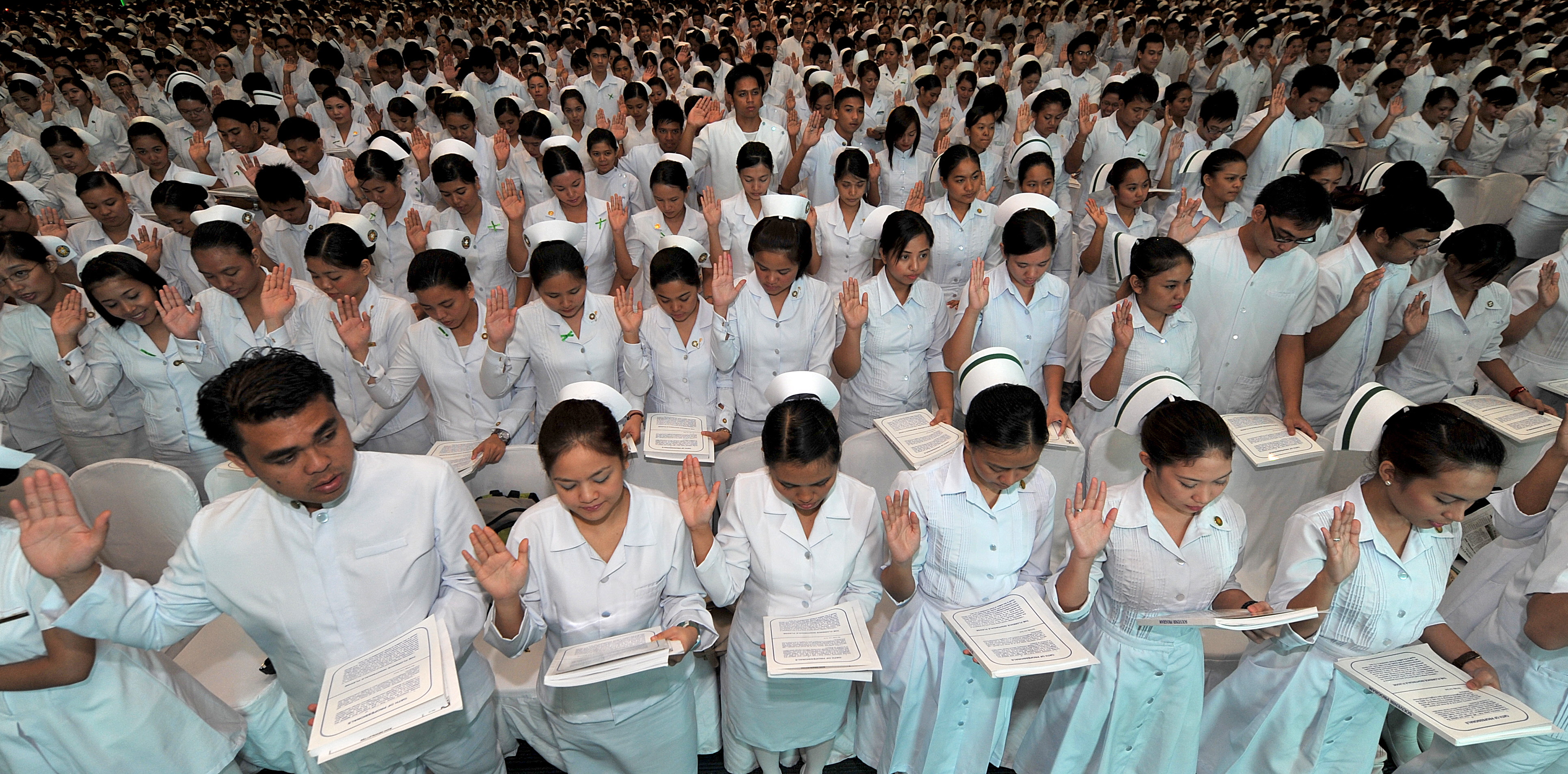 Women in rows wearing white nurses' uniforms and hats all have their right hand up, with their left holding a document they are reading from.