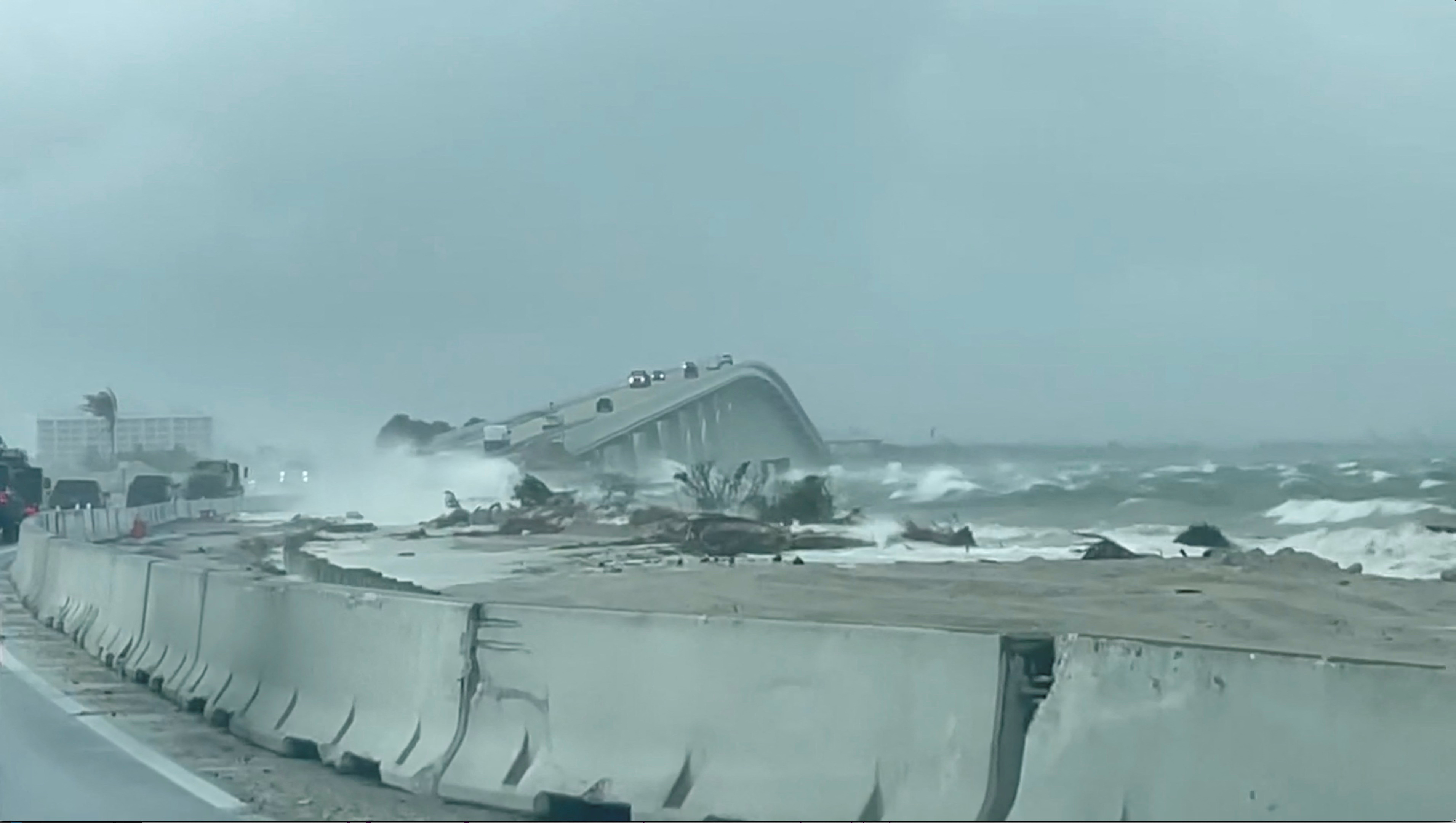 Waves and inundation batter a coastal bridge as cars attempt to cross.