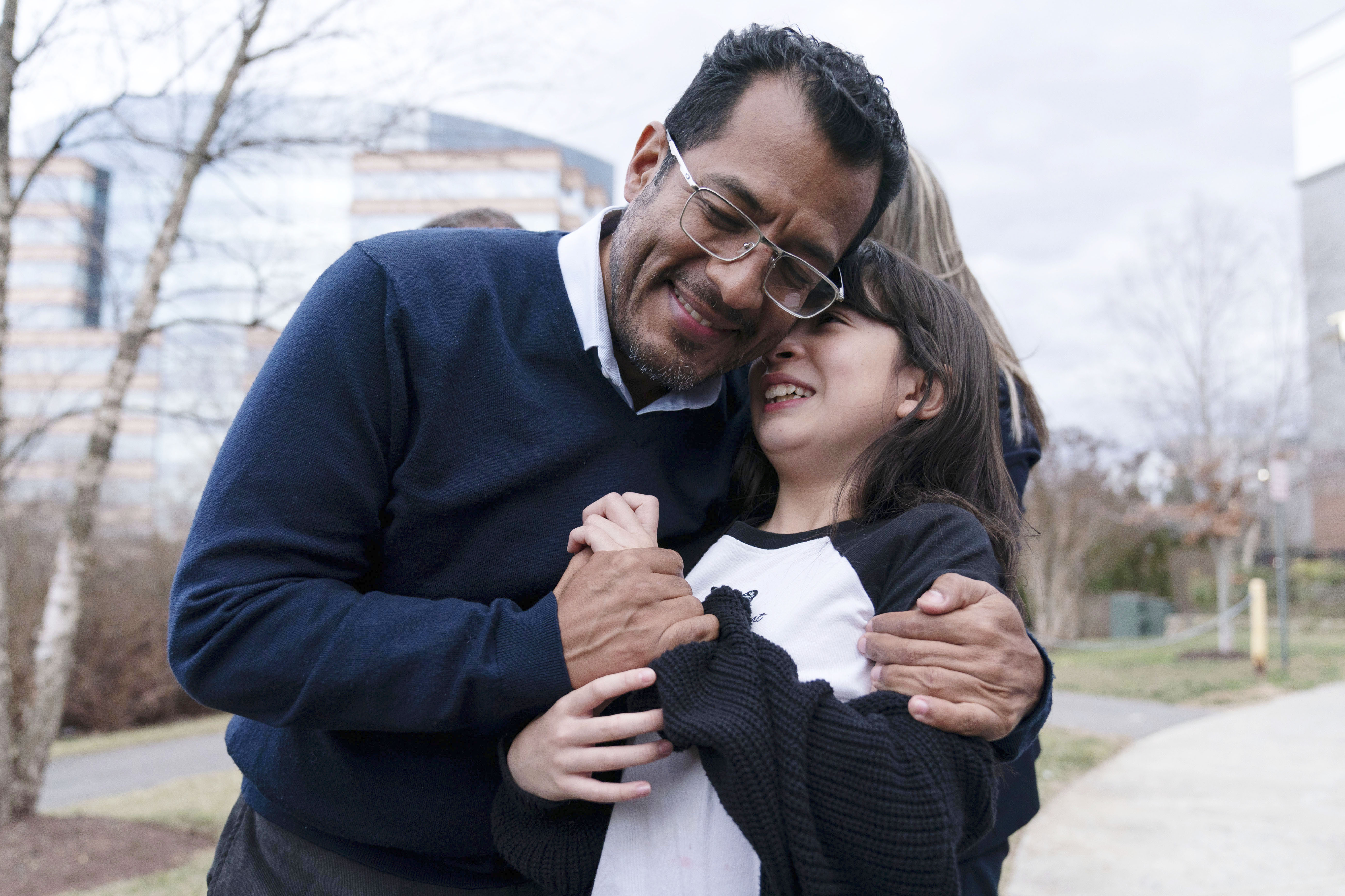 Former Nicaragua presidential candidate Felix Maradiaga hugs his young daughter Alejandra, outside on a sidewalk in the Washington, DC, area. She hides her face in his neck as he smiles and wraps his arms around her.