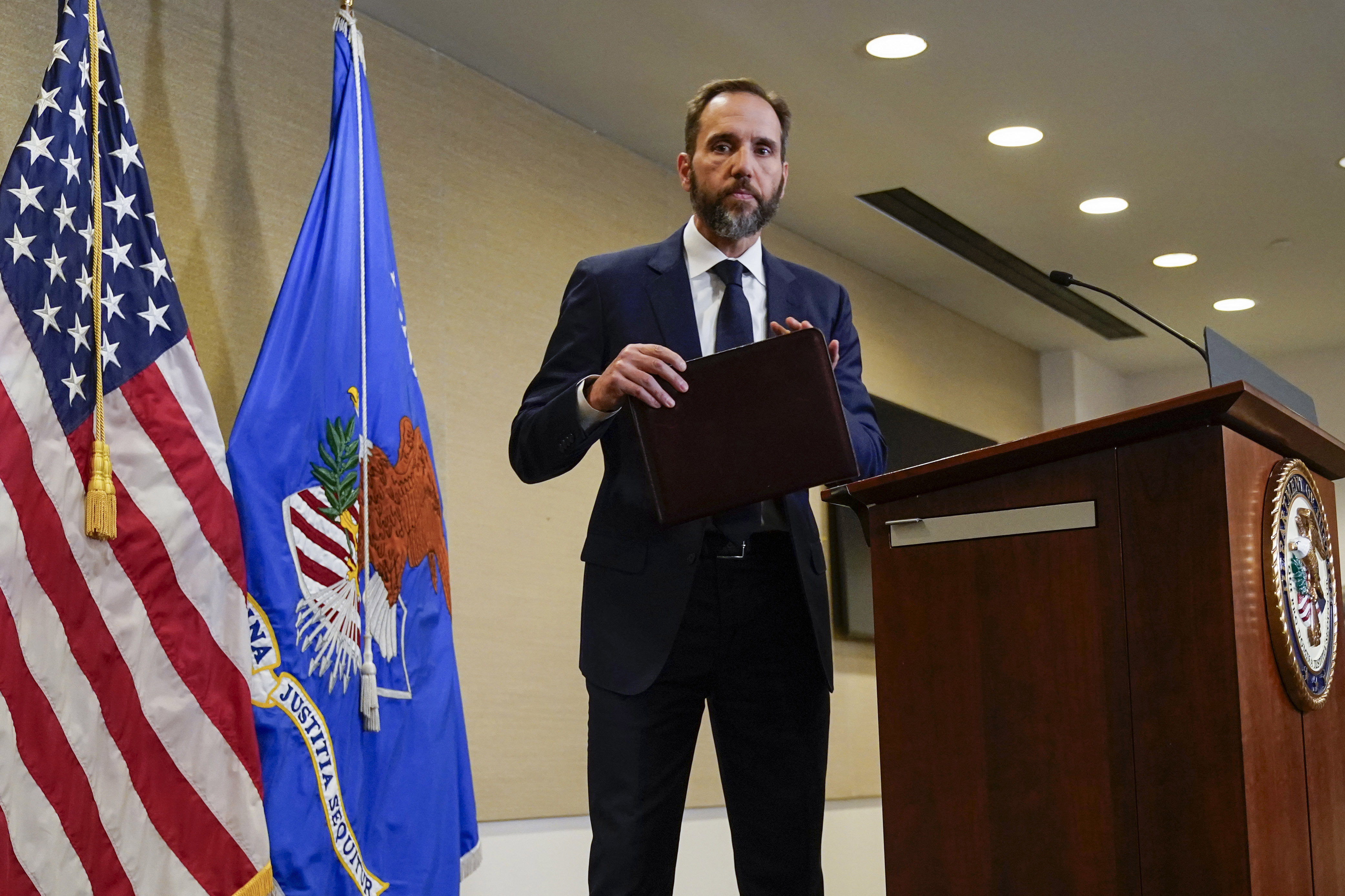 A man steps away from a podium, carrying a folder. Flags are behind him.