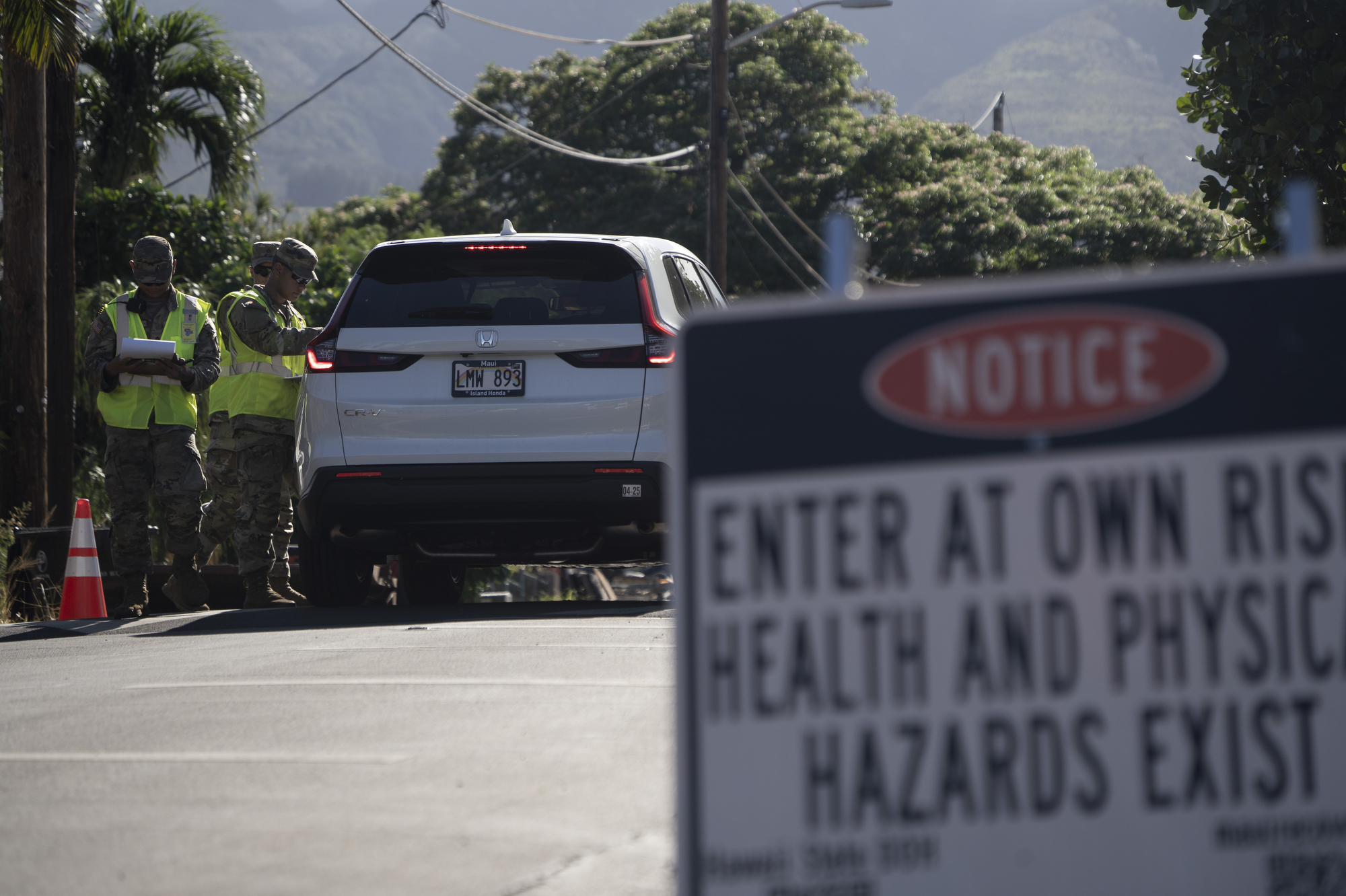 Two people in yellow traffic vests from the Hawaii National Guard stand outside a vehicle at a checkpoint. In the foreground, a sign reads, "Enter at your own risk. Health and physical hazards exist."