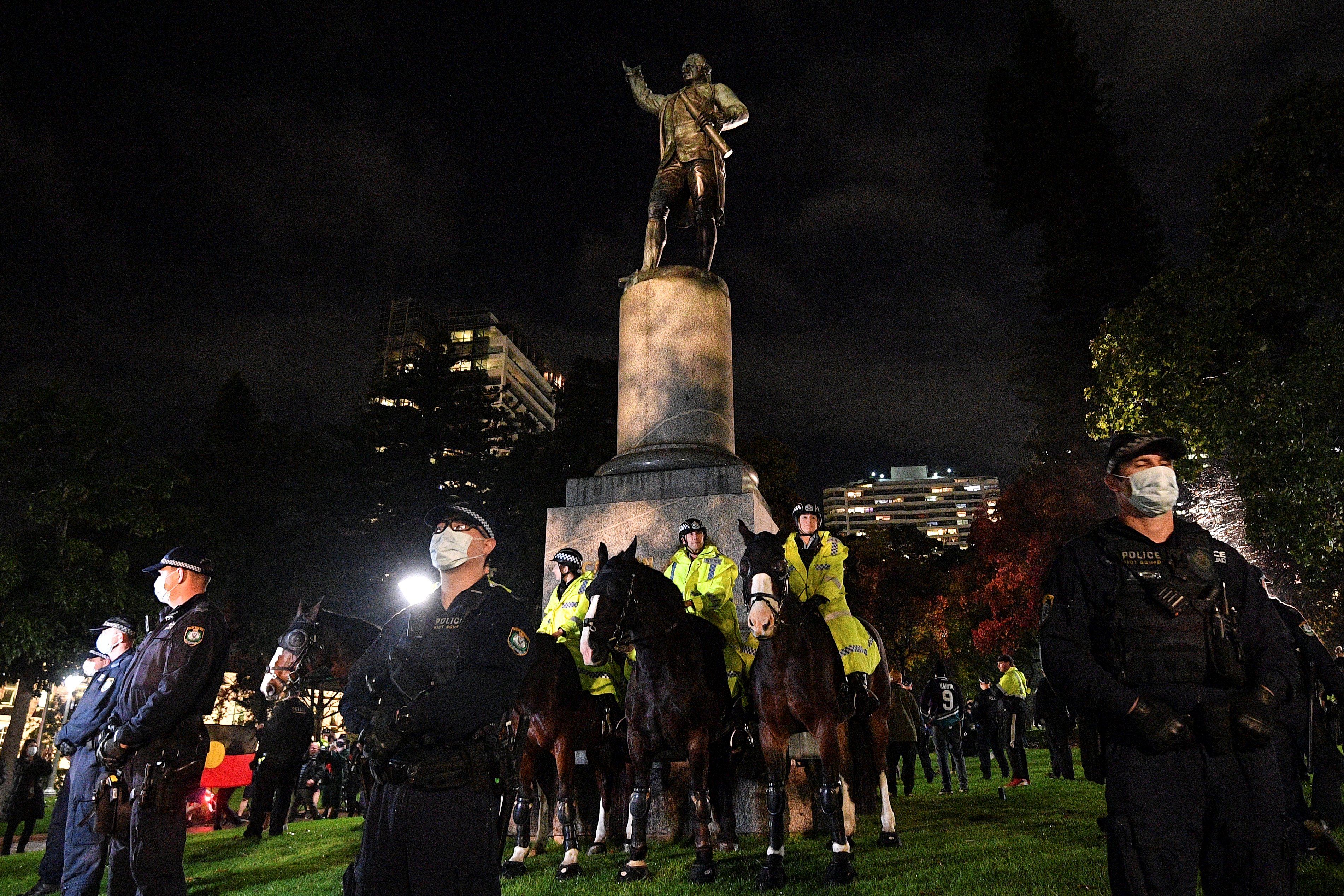 Three police in fluor yellow jackets sitting on horses in front of a statue of Captain Cook at night with a small glimpse of an Aboriginal flag
