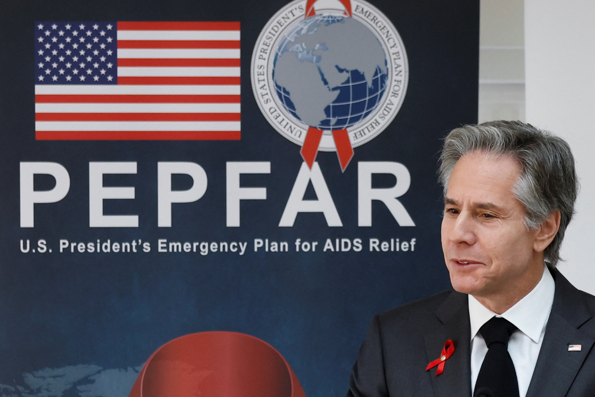 US Secretary of State Antony Blinken sits in front of a large blue banner for PEPFAR, with the acronym and the programme's logo emblazoned on it, alongside an American flag.