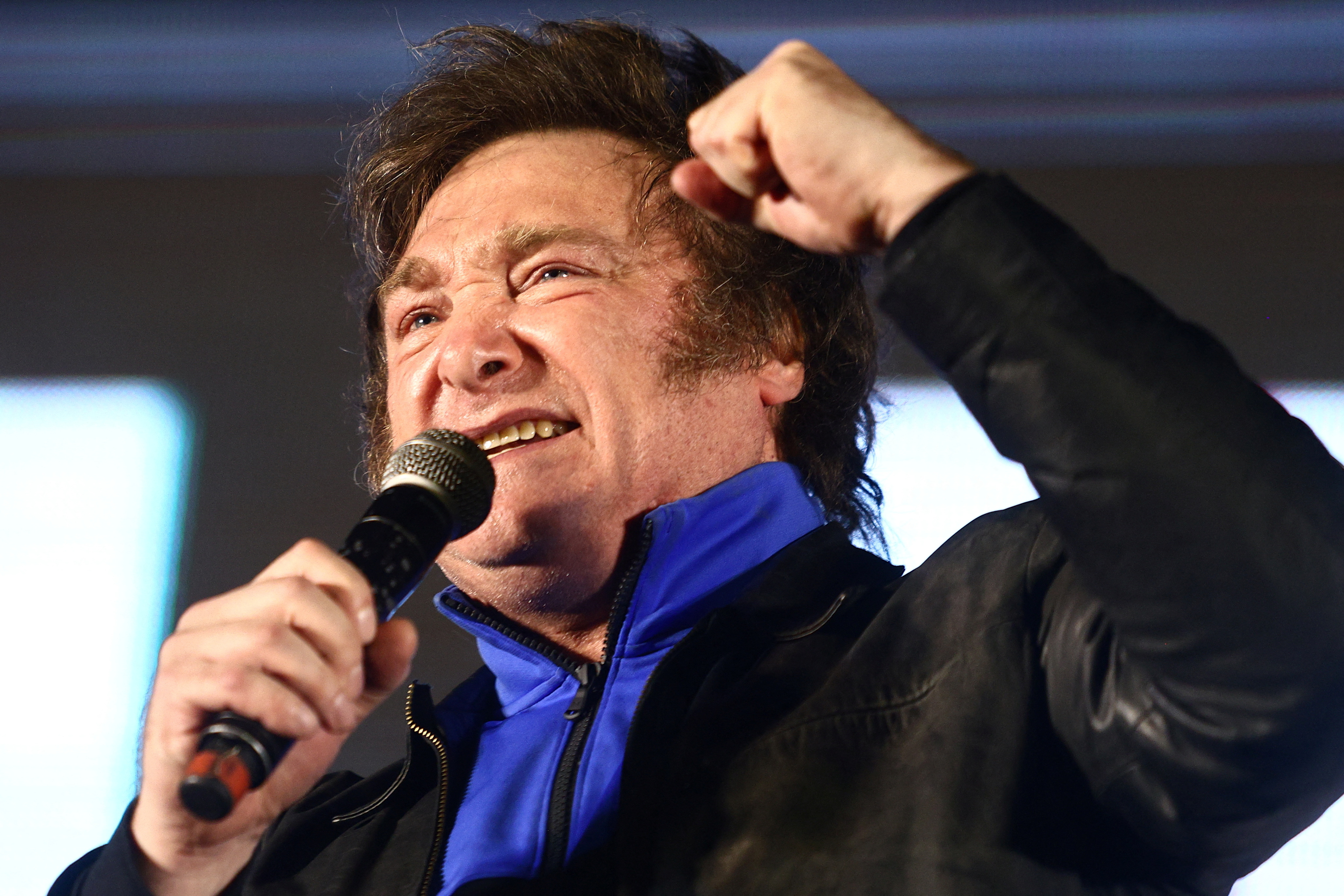 Javier Milei — dressed in a dark jacket and a blue sweater — raises a fist as he speaks into a microphone.