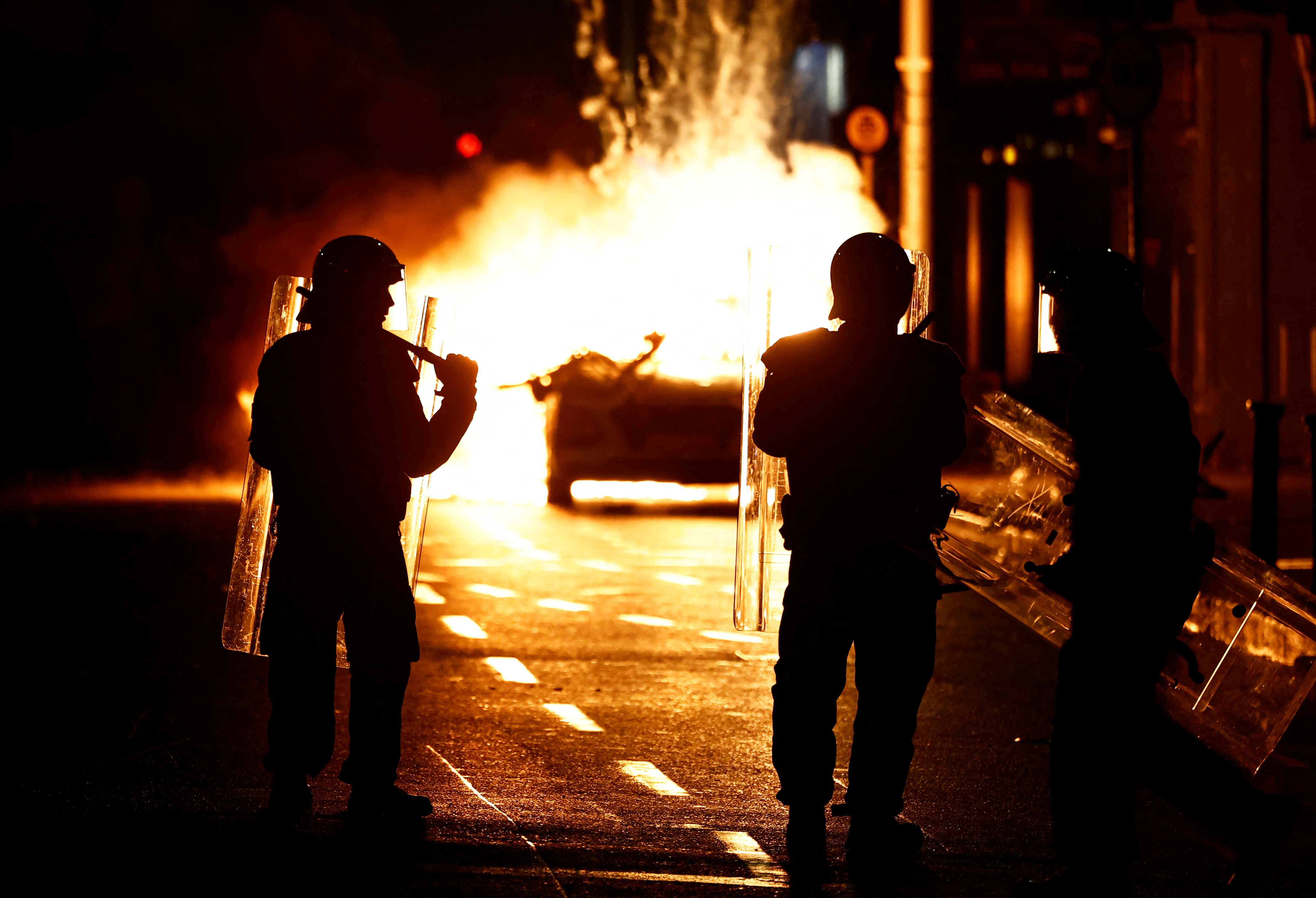 Riot police stands next to a burning police vehicle